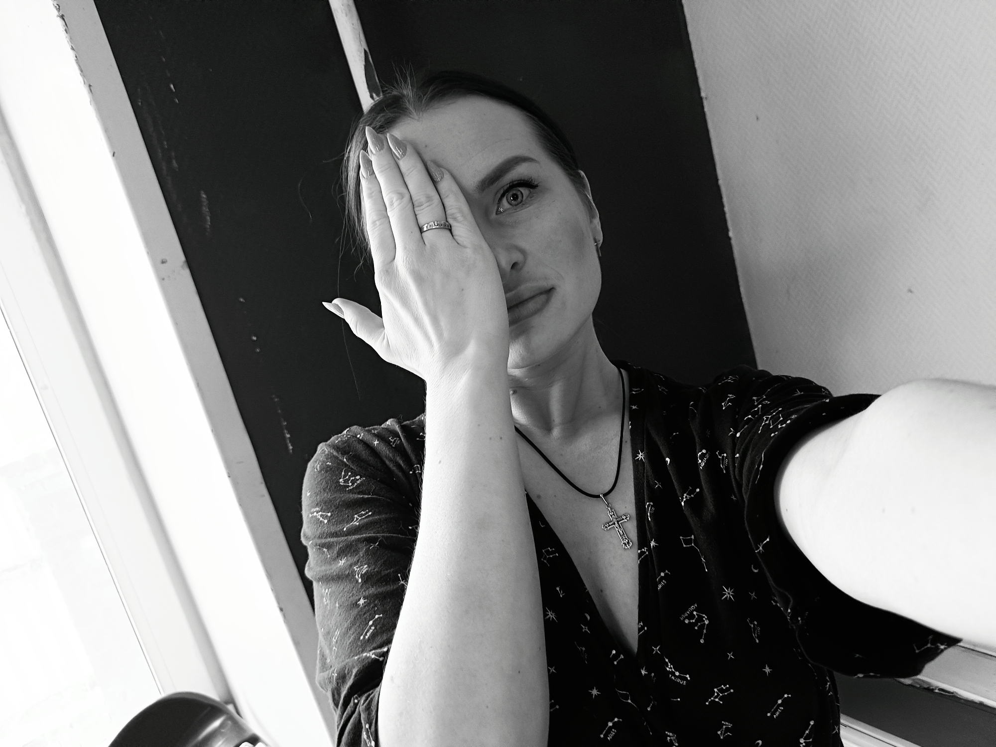 A woman is posing for a selfie indoors. She has one hand raised to cover half of her face. She has a silver ring on her ring finger. She is wearing a dark top with constellations printed on it, and a necklace with a cross pendant. The background includes a window frame and a wall. The image is in black and white. Rusalevka, Russia is not discernable from the image.
