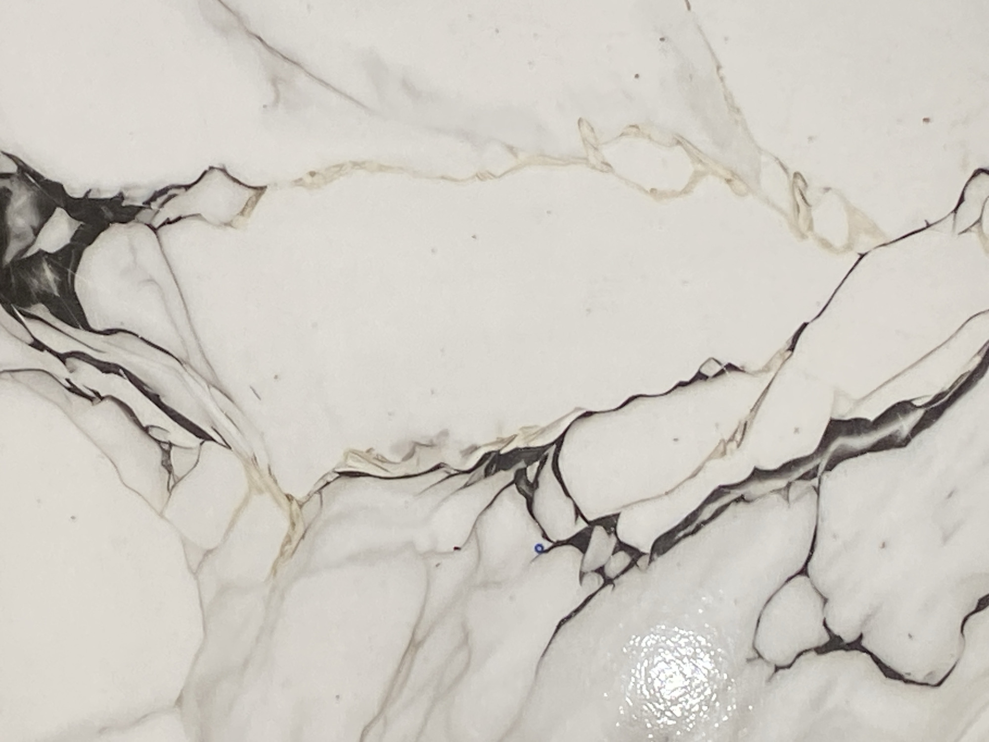 The image is a close-up, abstract view of a marbled surface. The dominant colors are white and shades of grey, with thin, irregular lines of golden-brown and black running through it, mimicking natural stone veining. There are no discernible people, objects, or a clear scene. The image focuses solely on the texture and pattern of the material, which appears to be ceramic tile or a similar engineered surface designed to look like marble. There are a few very small, scattered dark specks, possibly dust or imperfections on the surface. One very small blue dot is visible towards the center-right of the image, and a tiny red speck is visible to its left. The lighting suggests a neutral environment, possibly indoors, with some light reflection visible on the surface, indicating a smooth, potentially glossy finish. No text is visible. The location context of Jauro Yinu, Nigeria, does not offer specific cues visible within this abstract image.