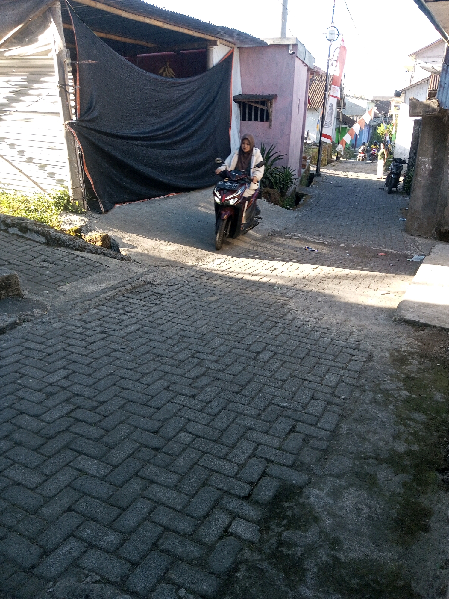 A street scene in Andongsili, Indonesia, shows a narrow paved lane between buildings. A woman wearing a hijab and a light-colored jacket rides a dark-colored motorcycle. The road is paved with interlocking grey bricks, and there are some patches of dirt and vegetation on the sides. Several Indonesian flags are visible, hanging from poles. In the background, other motorcycles and people can be seen. The buildings lining the street are small and appear to be residential or commercial establishments. The lighting suggests it is daytime, with shadows cast across the road.