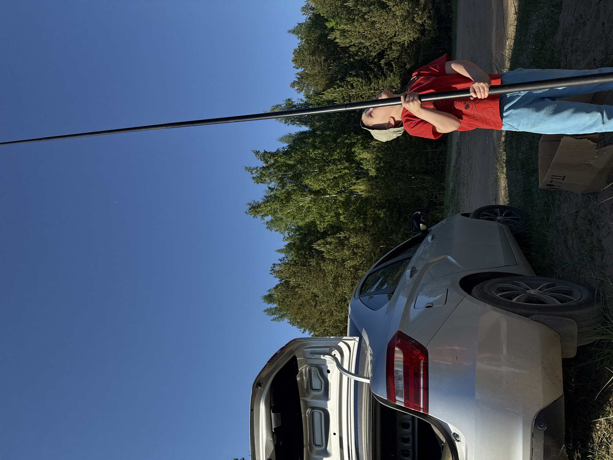 A photo taken outdoors, likely in a rural or suburban area, features a young person holding a long, black pole. The person is wearing a red t-shirt, blue pants, and a light-colored cap. They are standing next to a silver car with its trunk open. A cardboard box is on the ground nearby. The background consists of trees and a blue sky. The location is unknown and cannot be determined based solely on the visual information provided.