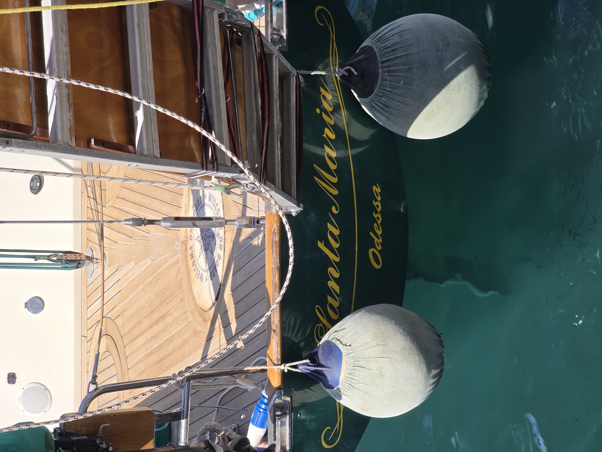 The image shows the stern of a dark green yacht named "Santa Maria" with the name "Odessa" written below it in yellow lettering. Two light gray fenders are hanging from the yacht, one on the left and one on the right. The yacht is docked in clear, turquoise water. The wooden deck of the yacht is visible at the top of the image, and a set of stairs is attached to the side of the yacht. The scene is lit by natural sunlight, casting shadows on the yacht and the water. The location is the Old Venetian Harbor in Chania, Crete, Greece.