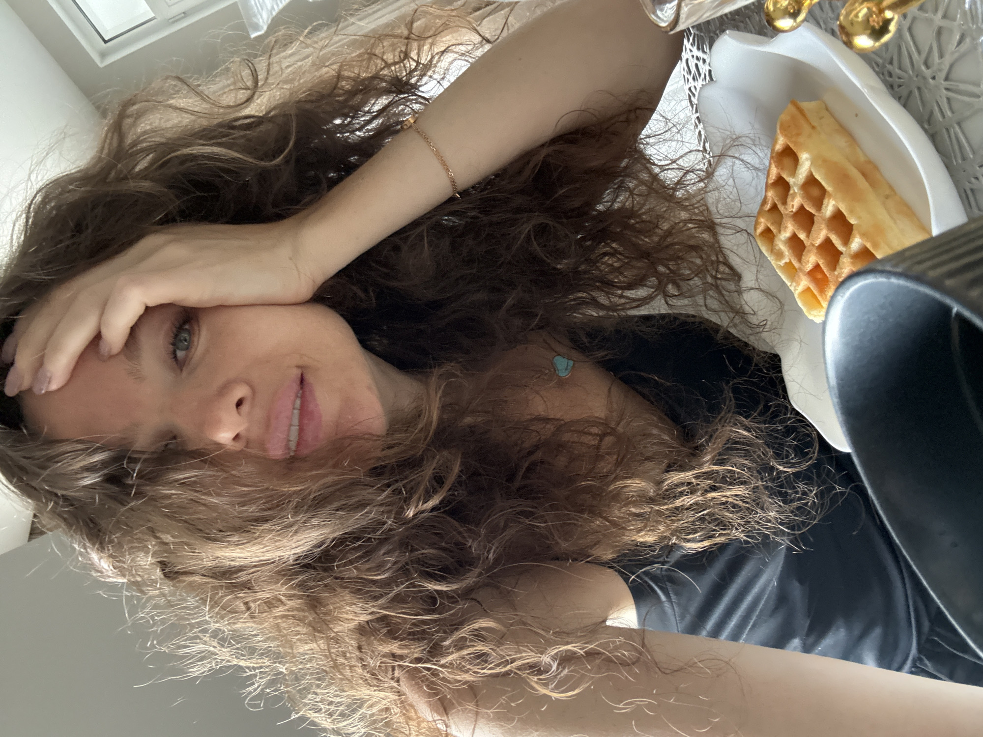 A woman with curly brown hair is positioned in the lower half of the image, partially covering her face with her right hand. She is wearing a black top and a gold bracelet on her left wrist. To the right of the woman, a white plate holds a waffle. A black object is partially visible on the right side of the plate. The location and city of Khimki, Russia are not discernable from the image.