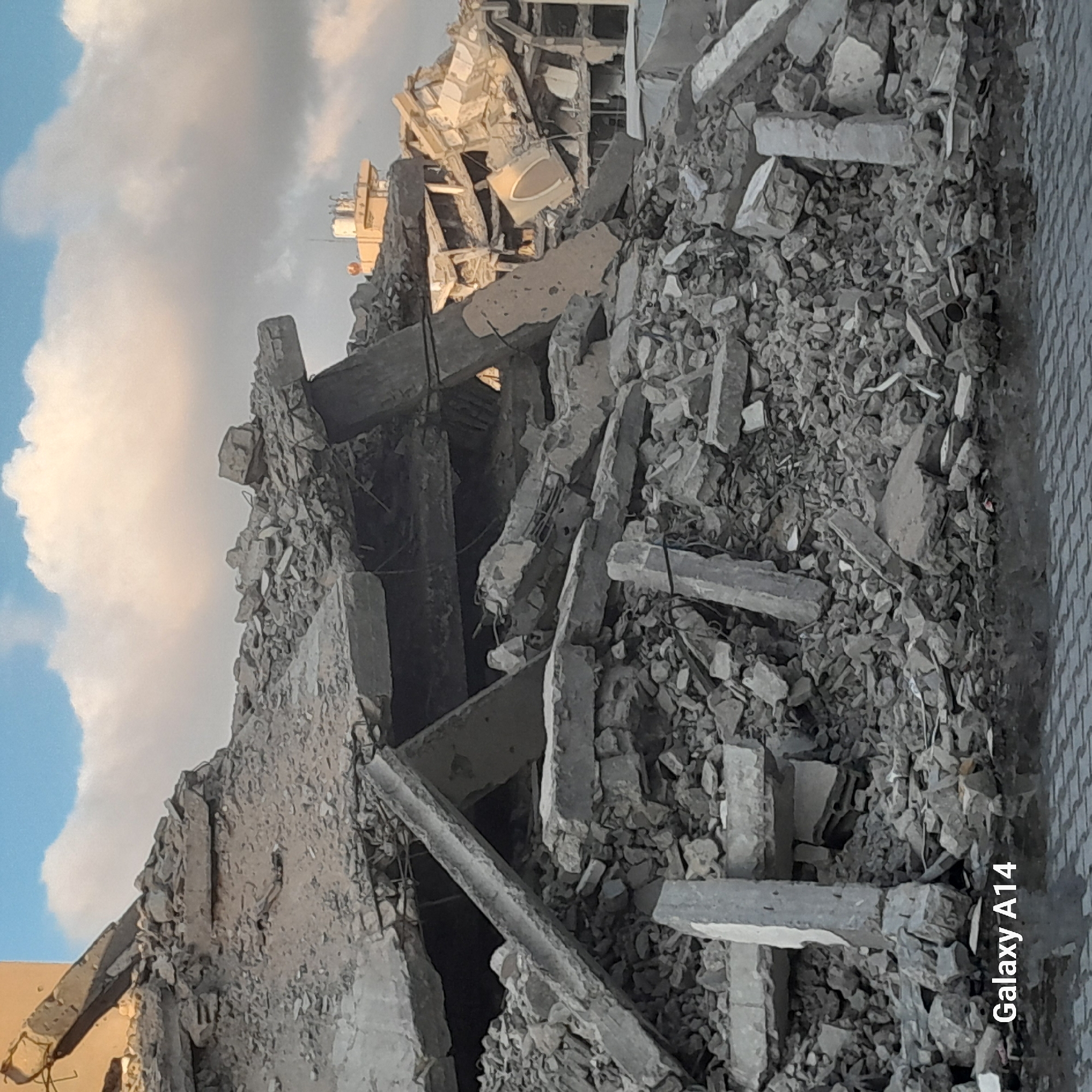 This image depicts the aftermath of destruction in the city of Khan Yunis, Palestinian Territories. The scene is dominated by a massive pile of rubble, consisting of concrete debris, twisted metal rebar, and shattered building materials. In the background, the skeletal remains of damaged structures are visible, some with exposed interiors. The sky is partly cloudy, with soft, diffused light illuminating the scene. In the foreground, the bottom of the image shows a reflective surface, possibly wet pavement or standing water, with a cobblestone pattern visible beneath. The words "Galaxy A14" are superimposed in white text in the lower left corner of the image. There are no people visible in the frame. The overall impression is one of devastation and ruin.
