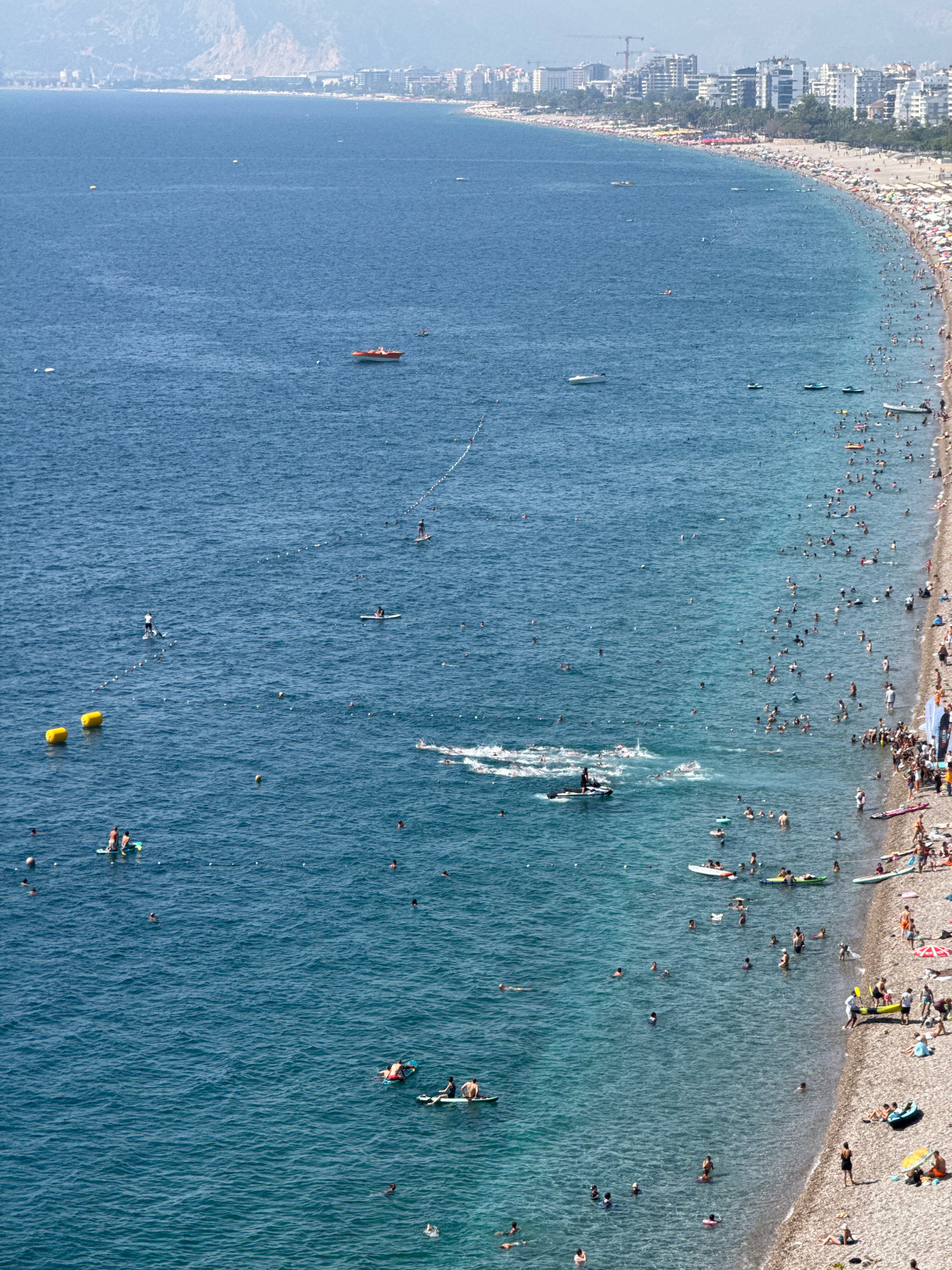 This is an aerial view of a crowded beach and the sea in Antalya, Turkey, likely in the Lara Beach area. The image shows a long stretch of coastline with a city skyline in the background. The sea is a clear blue, with numerous people engaged in various water activities.
In the water, there are many swimmers, some are in groups, while others are alone. Several individuals are on paddleboards, with some appearing to be standing and others sitting or lying down. There are also a few small boats and inflatable watercraft scattered across the sea. Yellow buoys mark a designated swimming area closer to the shore. Further out in the water, a red and white boat is visible.
The beach itself is densely populated with sunbathers relaxing on towels and under numerous umbrellas. People are walking along the shoreline, wading in the shallow water, and interacting with each other. Various beach equipment, such as chairs, towels, and a few water sports accessories like surfboards and kayaks, are visible on the sand. The coastline is lined with buildings, hotels, and palm trees, characteristic of a resort city. The overall impression is a vibrant and active beach scene on a sunny day.