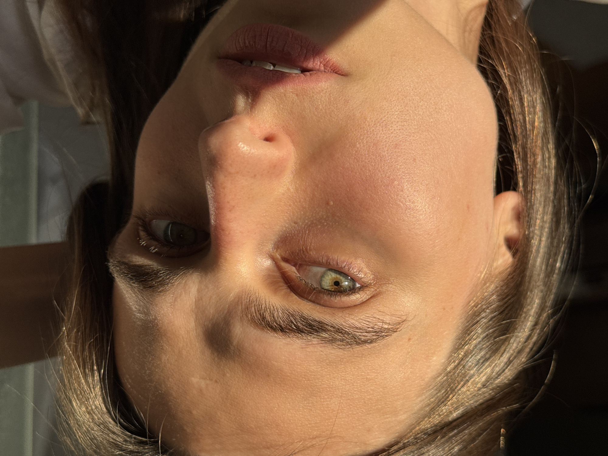 The image is a close-up, waist-up portrait of a young woman with light brown hair and green eyes. She is looking directly at the camera with a neutral expression. The lighting suggests it is either early morning or late afternoon, with sunlight casting a warm glow on her face and highlighting the texture of her skin. She appears to be indoors, possibly in a room with a window, as suggested by the shadow and the diffused light. There is no discernible background detail that would allow for specific identification of the location within Bečići, Montenegro. The overall impression is a natural and unposed photograph.