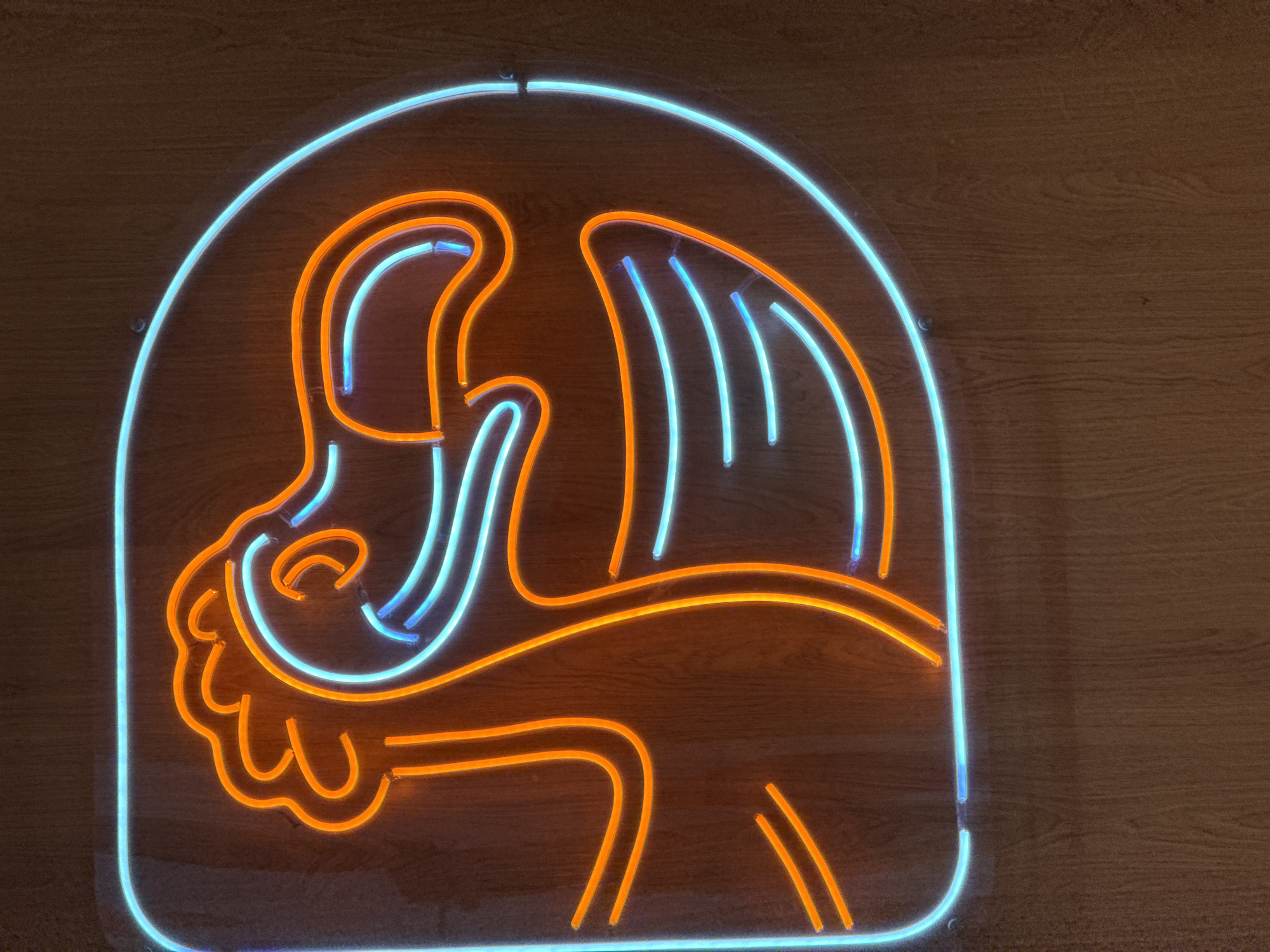 A neon sign depicting a lion in blue and orange, mounted on a wooden surface. The sign is composed of glowing neon tubes forming the outline of the lion's head and mane. The lion is set against a blue rounded square. The background is a wood-paneled wall. The location is Tobolsk, Russia.