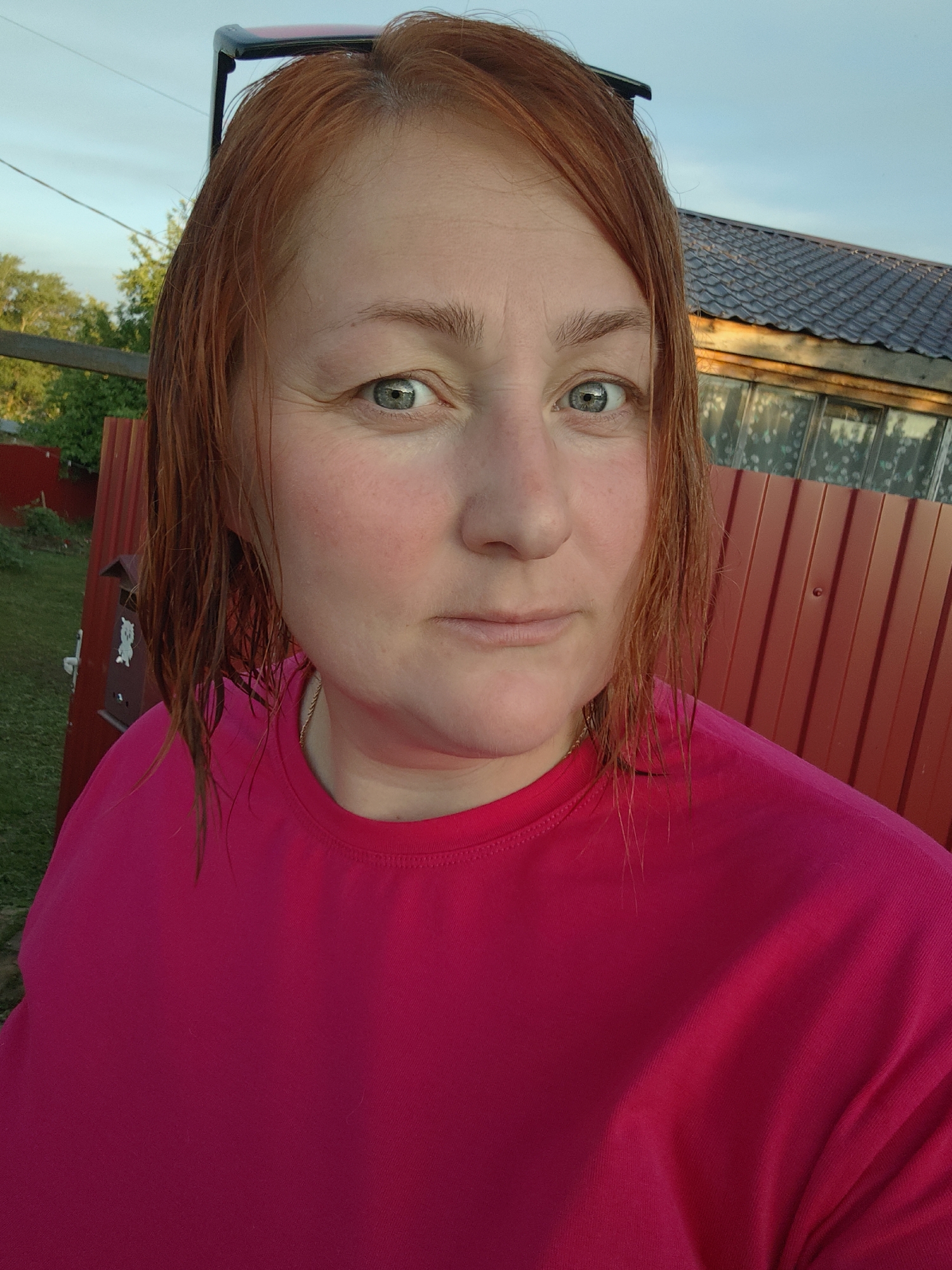 A close-up selfie captures a woman with reddish-brown, seemingly damp hair, looking directly at the camera with a neutral expression. Her eyes are a striking greenish-blue. She has some natural flush on her cheeks. She is wearing a bright fuchsia-pink short-sleeved t-shirt, and a thin gold chain is visible around her neck. Black-framed sunglasses rest on top of her head, partially obscured by her hair.
The background suggests an outdoor setting during late afternoon or early evening, characterized by warm, soft light. To her left, there's a red corrugated metal fence, and behind it, green foliage from trees and grass is visible. A small, light-colored decorative figure, possibly a cartoon animal, is attached to the fence. To her right, beyond the fence, a building with a dark, possibly tiled roof and multiple-paned windows is visible, suggesting a house or a shed/greenhouse. The sky above is clear and light blue. The overall scene indicates a rural or suburban environment, consistent with the location of Foky, Russia. There is no legible text visible in the image.