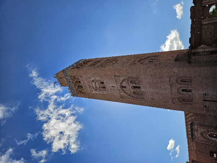 A tall, brown brick tower is seen angled upward against a bright blue sky with scattered white clouds. The tower features decorative arches and patterns along its height. The top of the tower has intricate designs and a distinct cornice. Part of an adjacent building with similar brickwork is visible to the right.