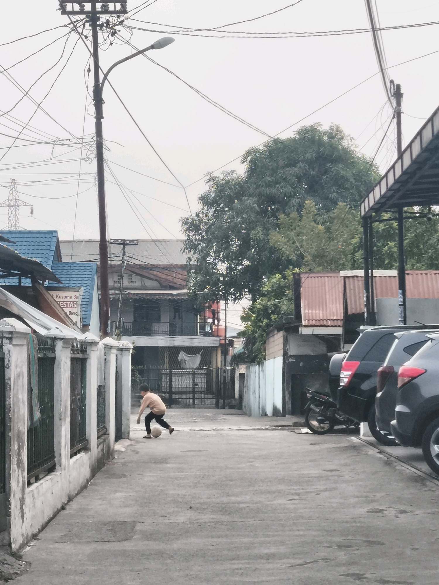 A young boy is playing soccer in a narrow alleyway in Palembang, Indonesia. The boy is wearing a light brown shirt and dark pants. He is kicking a white soccer ball. The alleyway is lined with houses and fences on the left and parked cars and a motorcycle on the right. Overhead, a tangle of electrical wires crisscrosses the sky, with a streetlamp attached to a utility pole. The houses are made of various materials, including corrugated metal and painted concrete. The overall scene is one of urban life in a densely populated area, with everyday activities taking place amidst the infrastructure.