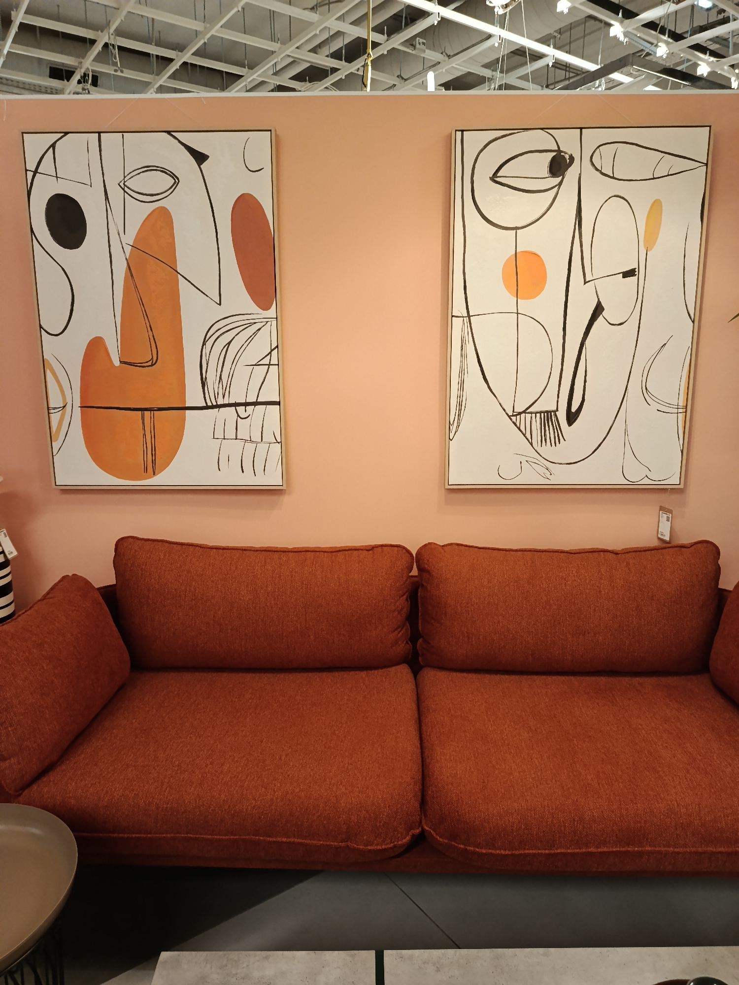 This media file presents an interior scene, likely a furniture showroom or a staged living area, situated in Moscow, Russia. The focal point is a vibrant display featuring a sofa and two abstract artworks against a soft, light peach-colored wall.
The wall is adorned with two large, framed abstract paintings. The left painting showcases prominent black lines, a black circle, a significant orange organic shape, and a reddish-brown form on a white background, evoking a stylized face. The right painting mirrors this style with black lines, an orange circle, a yellowish oval, and other abstract elements on a white canvas, also suggesting a facial composition.
Below these artworks is a substantial two-seater sofa, upholstered in a rich, textured rust-orange fabric. Its plush cushions suggest comfort and invite seating. On the left armrest, a partial view of a black and white striped object, possibly a cushion or vase, is visible.
The ceiling above is a white suspended grid system, characteristic of commercial or retail environments, with exposed metallic structures, wires, and various lighting fixtures. The floor, partially seen in the foreground, appears dark. Hints of other furniture include a round, light-colored table on the far left and a rectangular, light-colored table centered in the lower foreground.
No human activity is depicted; the scene is a static arrangement designed for display. A small white label with a barcode and indistinct text, likely product information or pricing, is affixed to the wall to the right of the sofa, near the right painting. The indoor setting with artificial lighting prevents any determination of the time of day or external weather conditions. The overall impression is one of modern interior design with a warm, artistic aesthetic.