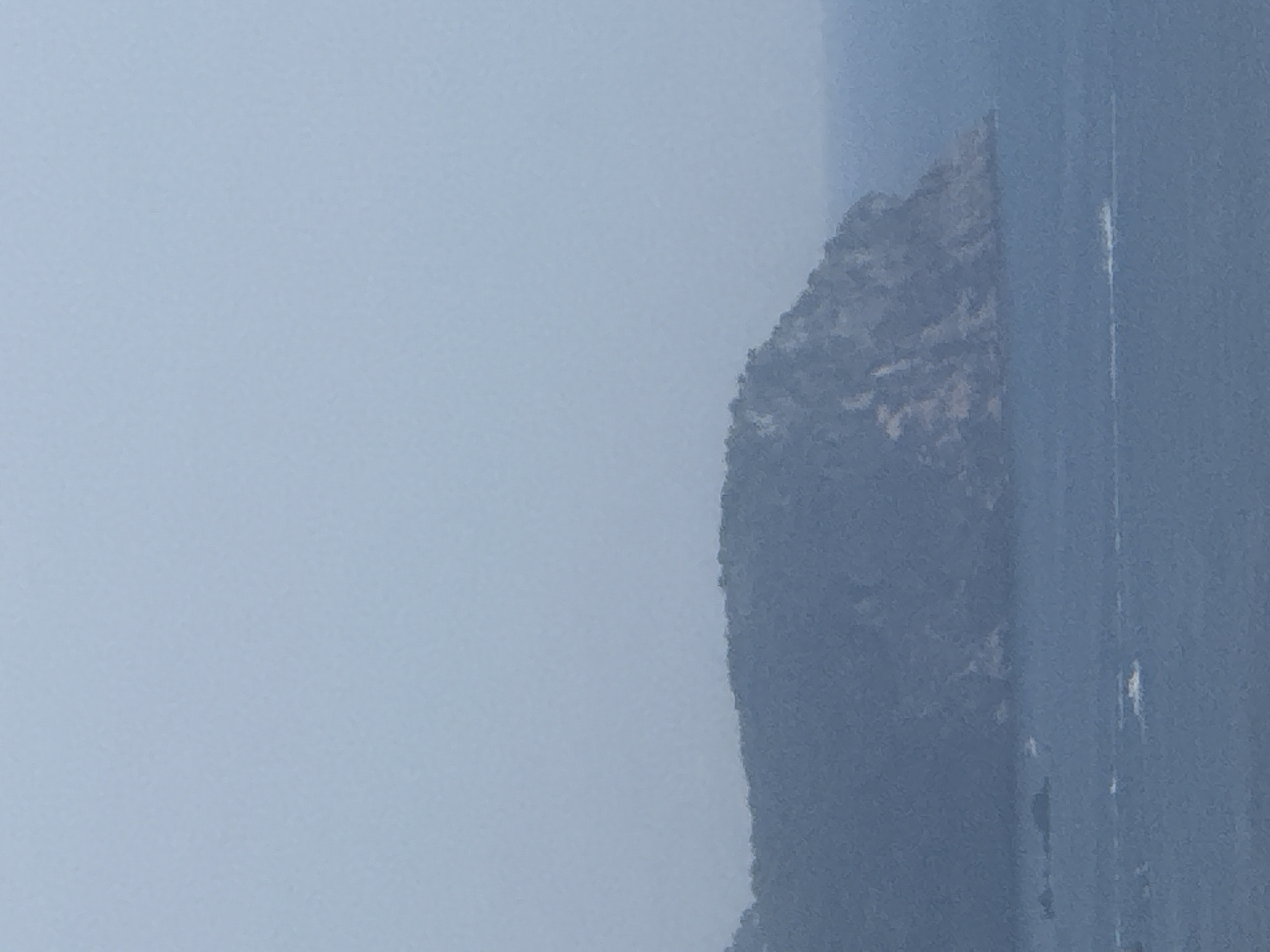 The image depicts a vertical, hazy landscape with a prominent, dark, mountainous feature dominating the right side of the frame. The mountain is steep and appears to be covered in dense vegetation, giving it a rough texture. To the right of the mountain, a vertical, lighter-colored streak, possibly a reflection or a thin waterfall, is visible. The background is a pale, uniform blue, suggesting either a very hazy atmosphere or the presence of fog.
The lighting is soft and diffused, indicating an overcast day or early morning/late evening. There are no visible people or man-made objects other than what might be suggested by the vertical streak. The overall impression is one of a remote, natural environment, possibly a fjord or a mountainous coastline given the presence of what might be water at the base of the mountain, though this is not clearly discernable. The image's orientation is vertical, and the lack of sharp detail due to the haze contributes to a sense of mystery and vastness. No text is visible in the image. The location context provided is Boreti, Montenegro, which aligns with the visual cues of a rugged, possibly coastal mountainous region.