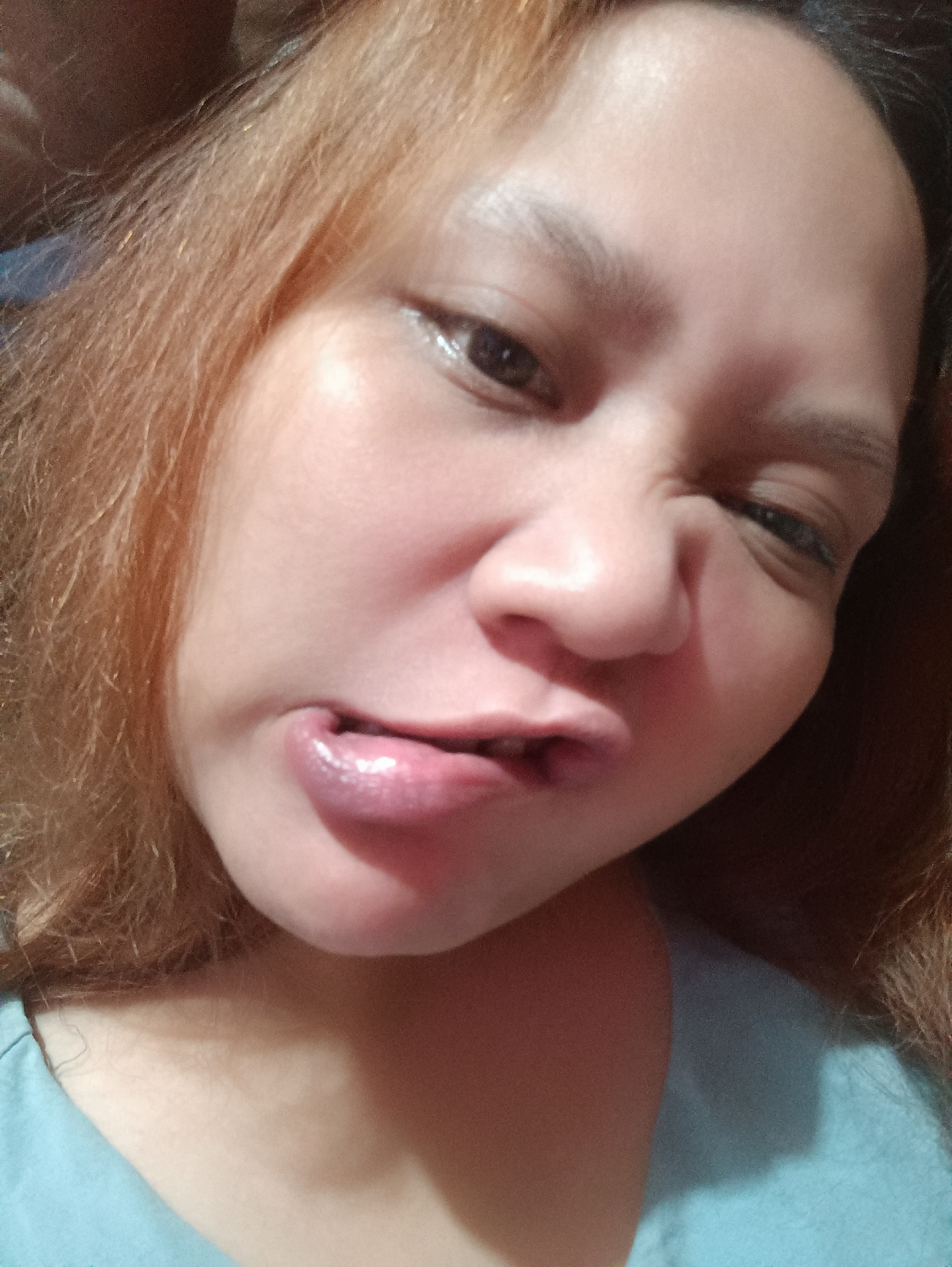 This is a close-up, low-angle, indoor selfie of an individual with light brown hair, making a grimacing facial expression. The individual is wearing a light blue shirt. The lighting is warm and appears to be artificial. The background is dark and out of focus, with no discernible features to indicate a specific location in Depok, Indonesia.