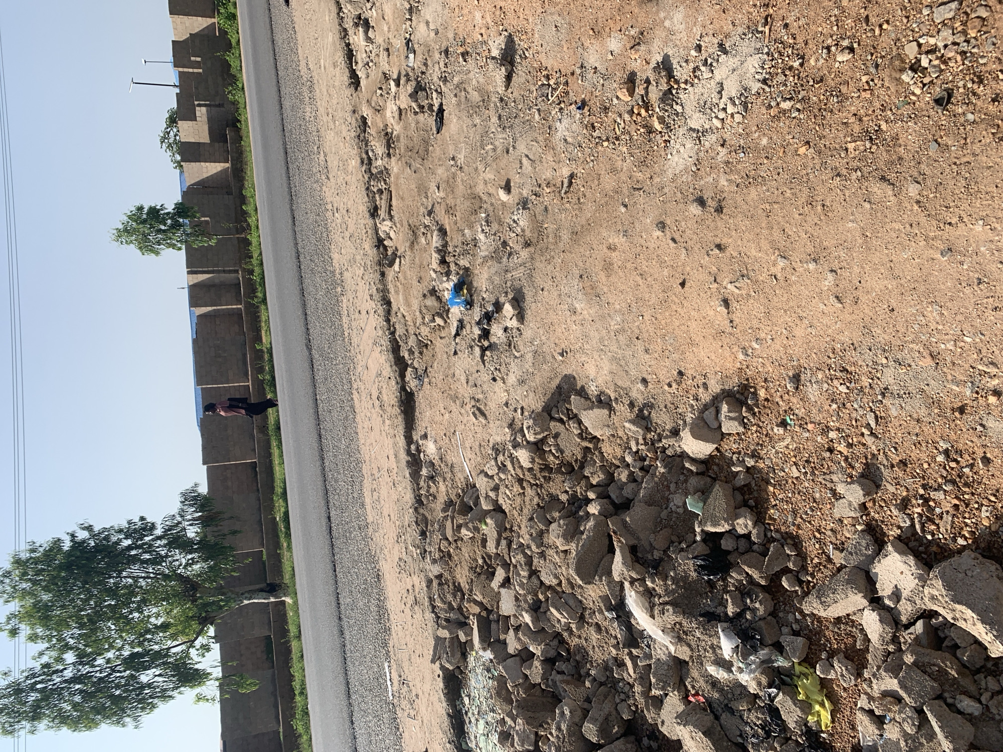 The image depicts a roadside in Jalingo, Nigeria, characterized by disarray and construction debris. In the foreground, a pile of broken concrete blocks and smaller stones lies on a dirt surface, interspersed with scattered plastic waste including a yellow plastic bag and a blue plastic bottle. A section of broken glass is also visible.
Further back, a paved road stretches horizontally across the frame. To the left of the road, a person in a dark shirt and pants, carrying a bag, walks away from the camera. Beside them, a large tree with green foliage stands, its trunk light-colored. A brick wall, constructed from reddish-brown bricks, runs along the edge of the road, punctuated by sections of lighter colored material and interspersed with smaller trees and vegetation. Two lampposts are visible in the distance, beyond the wall. The sky above is clear and pale blue. The overall impression is one of an undeveloped or under-construction area bordering a paved thoroughfare.