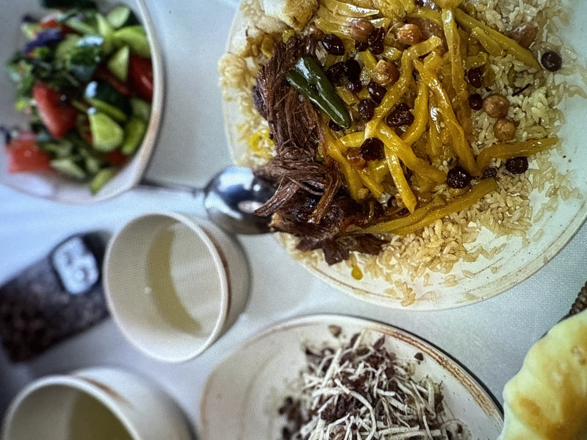 This is a top-down, close-up shot of a meal set on a light-colored tablecloth. The main focus is a plate of food in the upper right quadrant, featuring fluffy rice topped with shredded beef, long yellow strips that appear to be cooked carrots or another vegetable, and dark, dried fruits that could be raisins or dates. A single green chili pepper rests on top of the beef. To the left of this dish, a bowl of salad is visible, containing chopped tomatoes, cucumbers, and leafy greens.
In the lower half of the image, there are several small, cream-colored bowls, one of which is filled with a light yellow liquid. Another bowl in the bottom center contains a pile of shredded brown and white ingredients, possibly a topping or condiment. A dark, cylindrical object with a white symbol on it is partially visible in the lower left corner, suggesting it might be a spice grinder or a container. The lighting suggests it could be daytime, indoors. There is no visible text in the image. The overall scene suggests a leisurely meal being enjoyed.