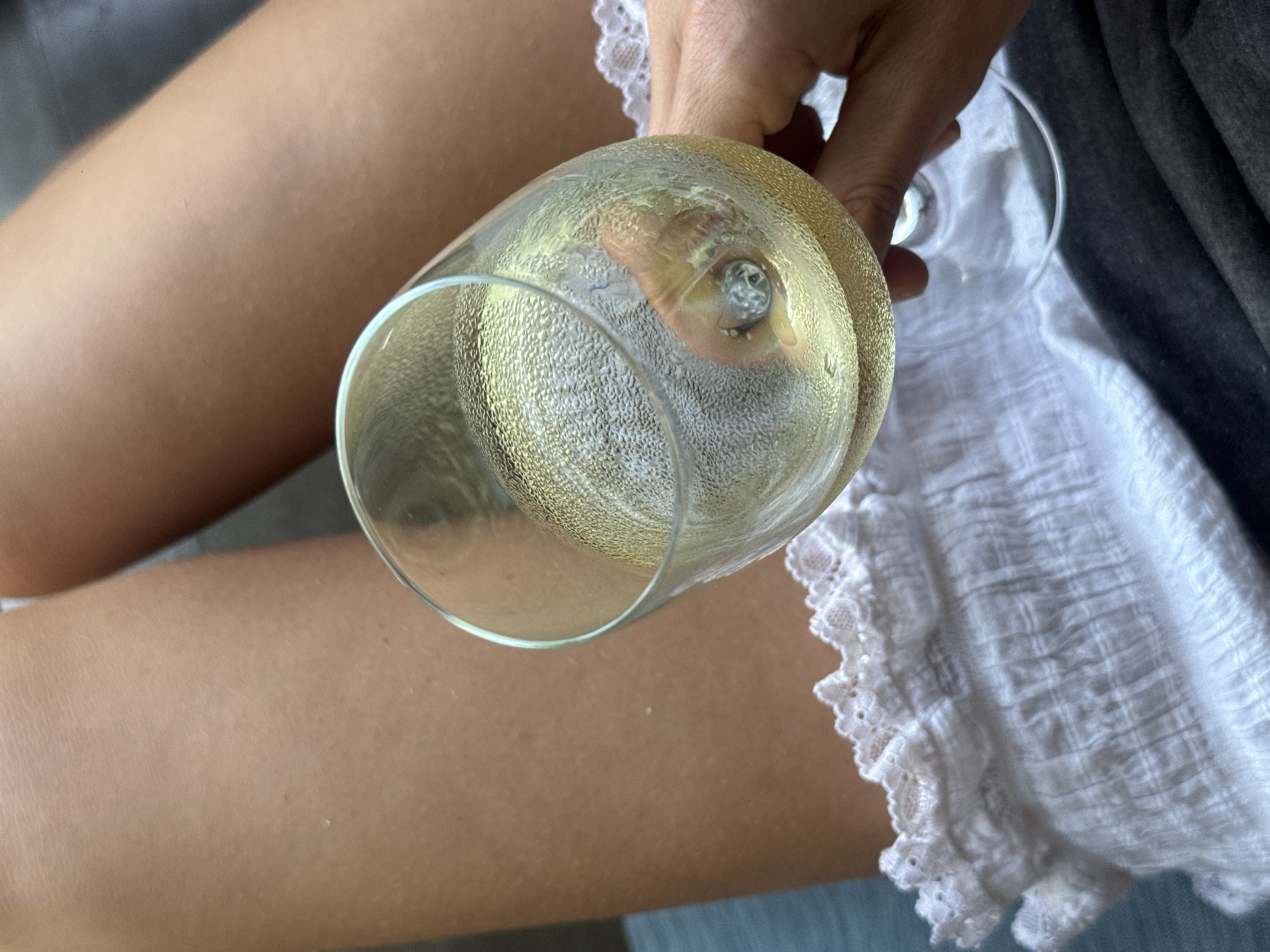 A person is holding a partially filled wine glass. The glass appears to contain a light-colored liquid, possibly white wine or champagne. The person's hand is visible, holding the base of the glass. Parts of the person's legs, a white lacy garment, and a grey garment are also visible in the frame.
The image's location is unspecified as North Kuta, Indonesia.