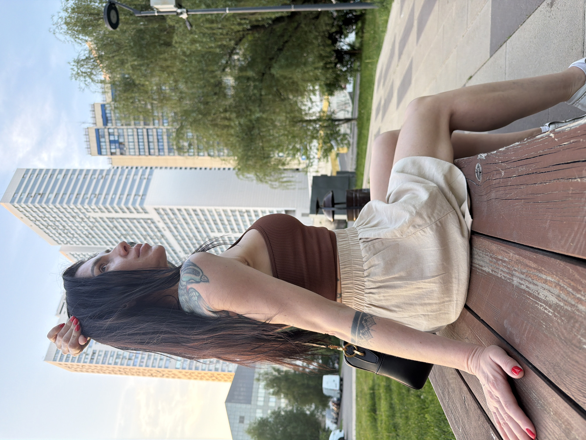 A woman is lying on a wooden bench outdoors. She is wearing a brown top and beige shorts. She has long, dark hair and tattoos on her arms. Her fingernails are painted red. She is looking up at the sky. A black purse is slung over her shoulder and rests on the bench. In the background, there are several tall buildings and green trees. This image was taken in Moscow, Russia.