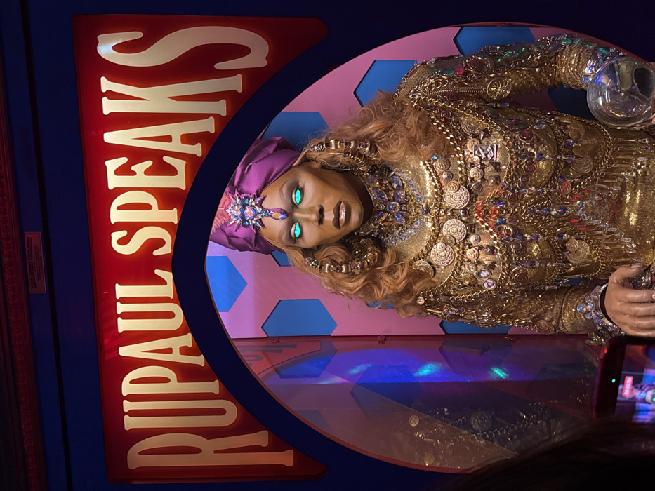 A wax figure of RuPaul dressed as a fortune teller, with glowing green eyes, is displayed. She wears a bejeweled turban and is adorned with gold clothing and accessories. The figure holds a crystal ball. A red sign that says “RuPaul Speaks” is situated to her left. The display case has a pink, blue, and purple background. The photo is slightly tilted.