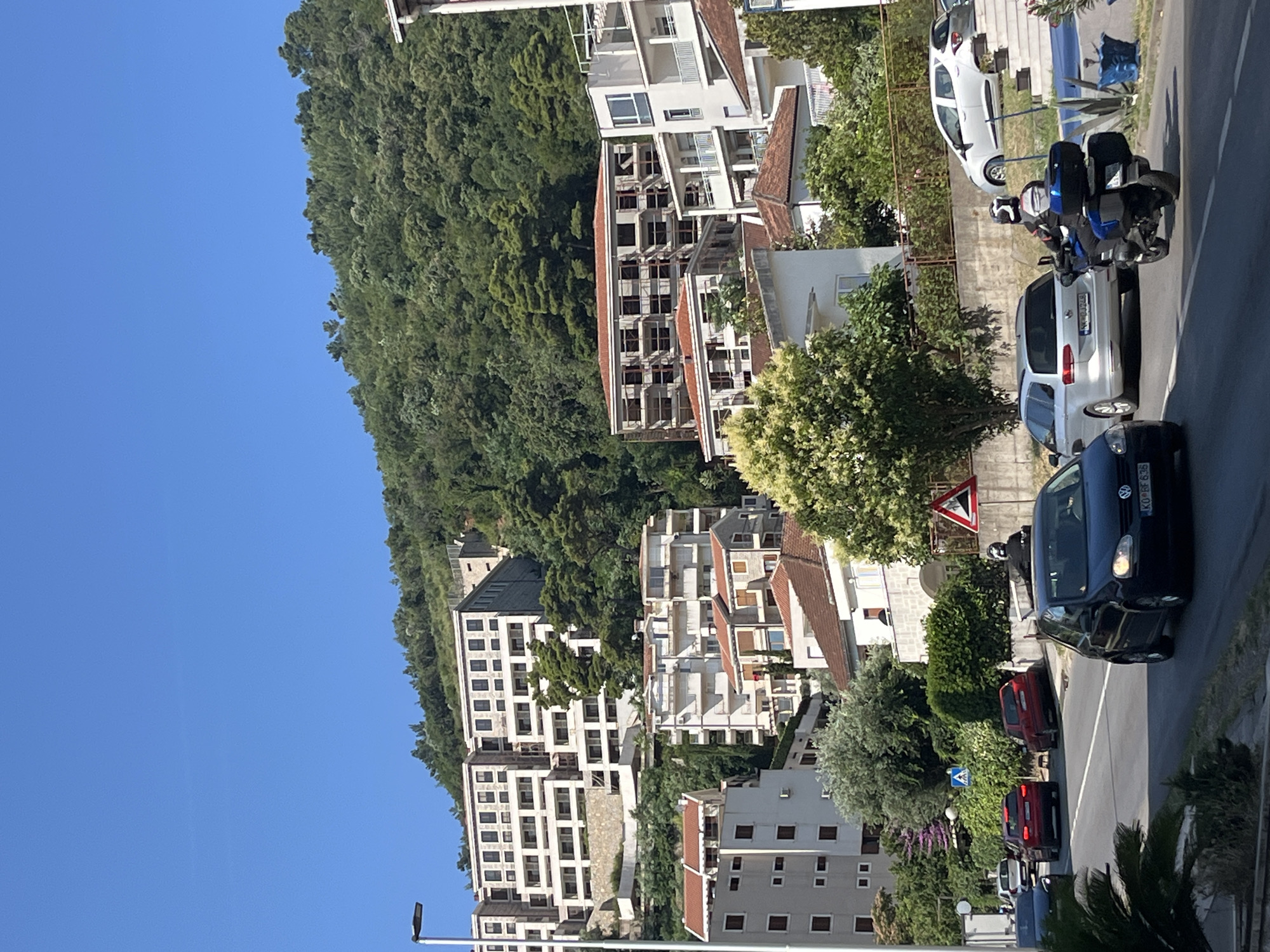 The image captures a sun-drenched street scene in Budva, Montenegro, with a focus on the hilly landscape dotted with buildings and dense greenery. In the foreground, a dark blue Volkswagen Golf is driving on the right side of the road, its headlights on. A motorcyclist wearing a helmet and protective gear is riding a blue motorcycle behind the Golf. To the left of the Golf, two red cars are visible on the road, one in front of the other. Further back, a silver station wagon and a white hatchback are parked on an elevated area to the right, near a set of stairs.
The mid-ground features a cluster of residential buildings with white facades and red-tiled roofs, nestled amongst lush green trees. One building appears to be under construction, with exposed concrete framing. The background is dominated by a steep, heavily wooded hillside, with more buildings peeking through the trees as they ascend the slope. The sky is a clear, vibrant blue. The overall impression is of a Mediterranean coastal town with its characteristic architecture and abundant vegetation.