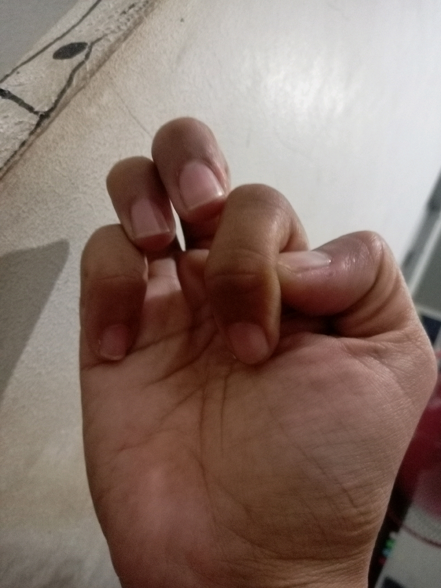 The image shows a close-up of a person's hand, with the fingers curled inwards. The hand is positioned against a textured, light-colored surface, possibly a wall or a piece of fabric, which has a dark, abstract pattern on the upper left side. The lighting suggests an indoor setting, casting a shadow on the textured surface. The skin tone of the hand appears to be medium brown. No specific location in Batam, Indonesia, can be identified from the image.