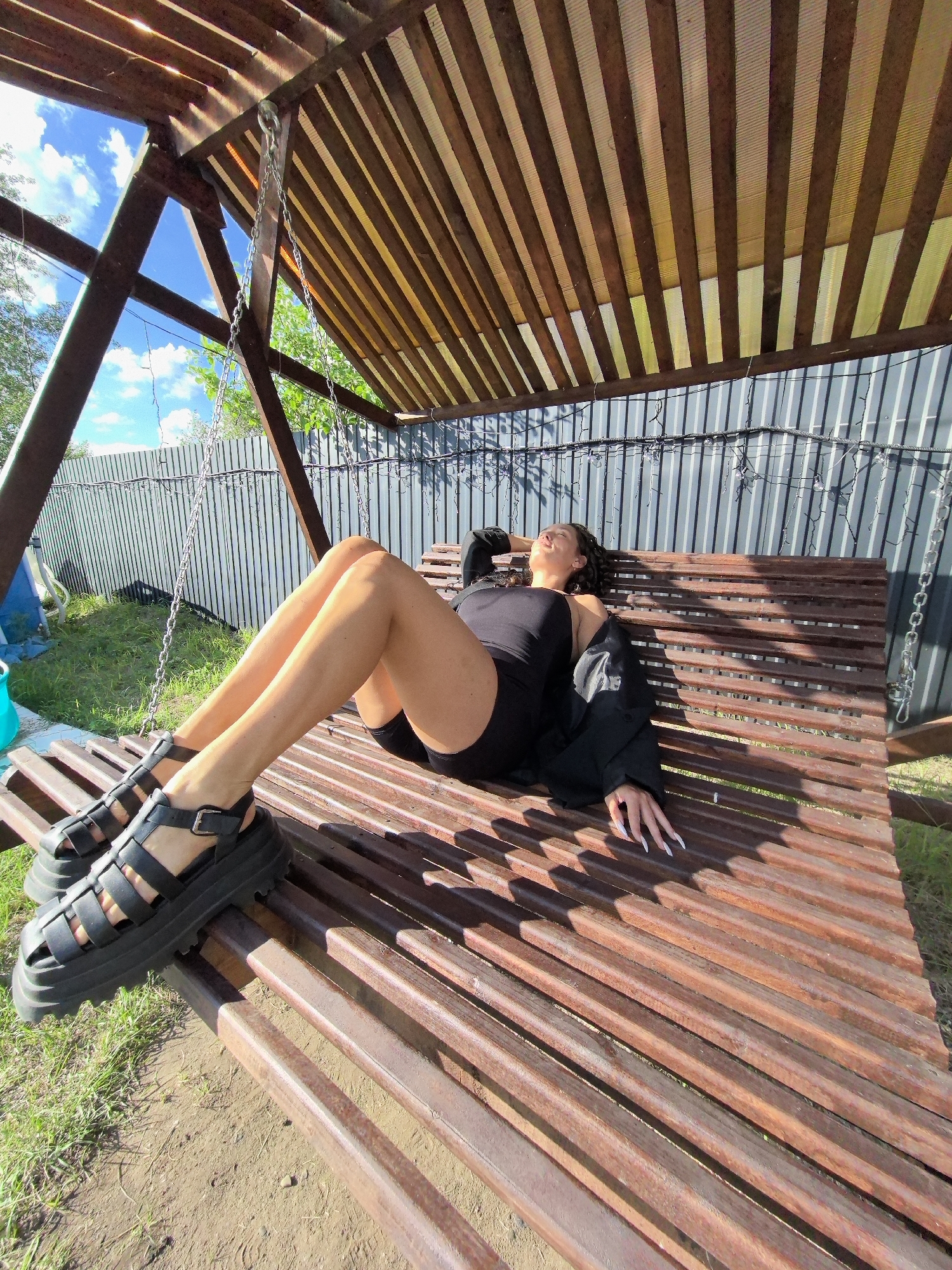 A young woman with dark curly hair is lying on a wooden swing bench in an outdoor setting. She is wearing a black sleeveless top, black shorts, a black jacket draped over her shoulders, and black platform sandals. Her right arm is resting behind her head, and her eyes are closed. The swing is made of wooden slats and is suspended by metal chains from a wooden structure. Behind the swing is a grey corrugated metal fence. The ground in front of the swing is dirt and grass. The sky above is blue with scattered white clouds, and there are trees and greenery visible in the background. The lighting suggests it is daytime, with strong sunlight casting shadows. The scene appears to be a backyard or garden area.