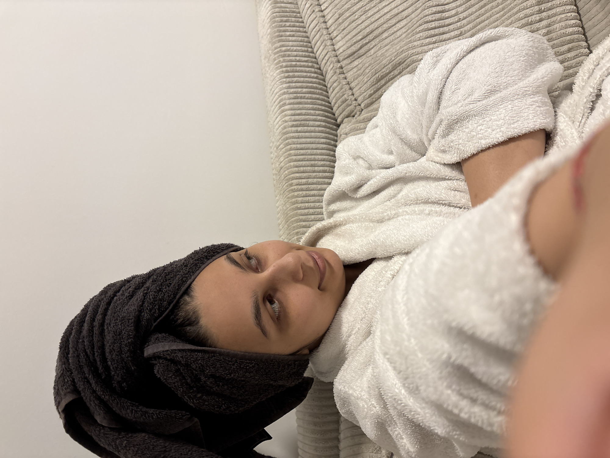 This close-up image captures a young adult, likely female, relaxing indoors. The person is wearing a white, fluffy bathrobe and has a dark grey towel wrapped around their head, suggesting they have just taken a shower or bath. They are reclining on a light grey or beige ribbed surface, possibly a sofa or a cushioned bed headboard, which occupies the right side of the frame. A plain, off-white wall forms the background on the left.
The subject's face is visible, turned slightly upwards and to the right, with their eyes open and looking away from the camera. A calm and serene expression is visible, hinting at a moment of peace and comfort. Part of a limb, out of focus and covered by the white bathrobe, is visible in the extreme foreground on the right, indicating a very close-up shot.
The setting is an indoor space, likely a living room or bedroom. The overall color palette is dominated by neutral tones: white, light grey/beige, dark grey, and natural skin tones. There are no specific external location cues visible in the image itself, though the provided context places the location in Mönchengladbach, Germany. No visible text is present. The soft, even lighting suggests an indoor light source and contributes to the relaxed atmosphere.