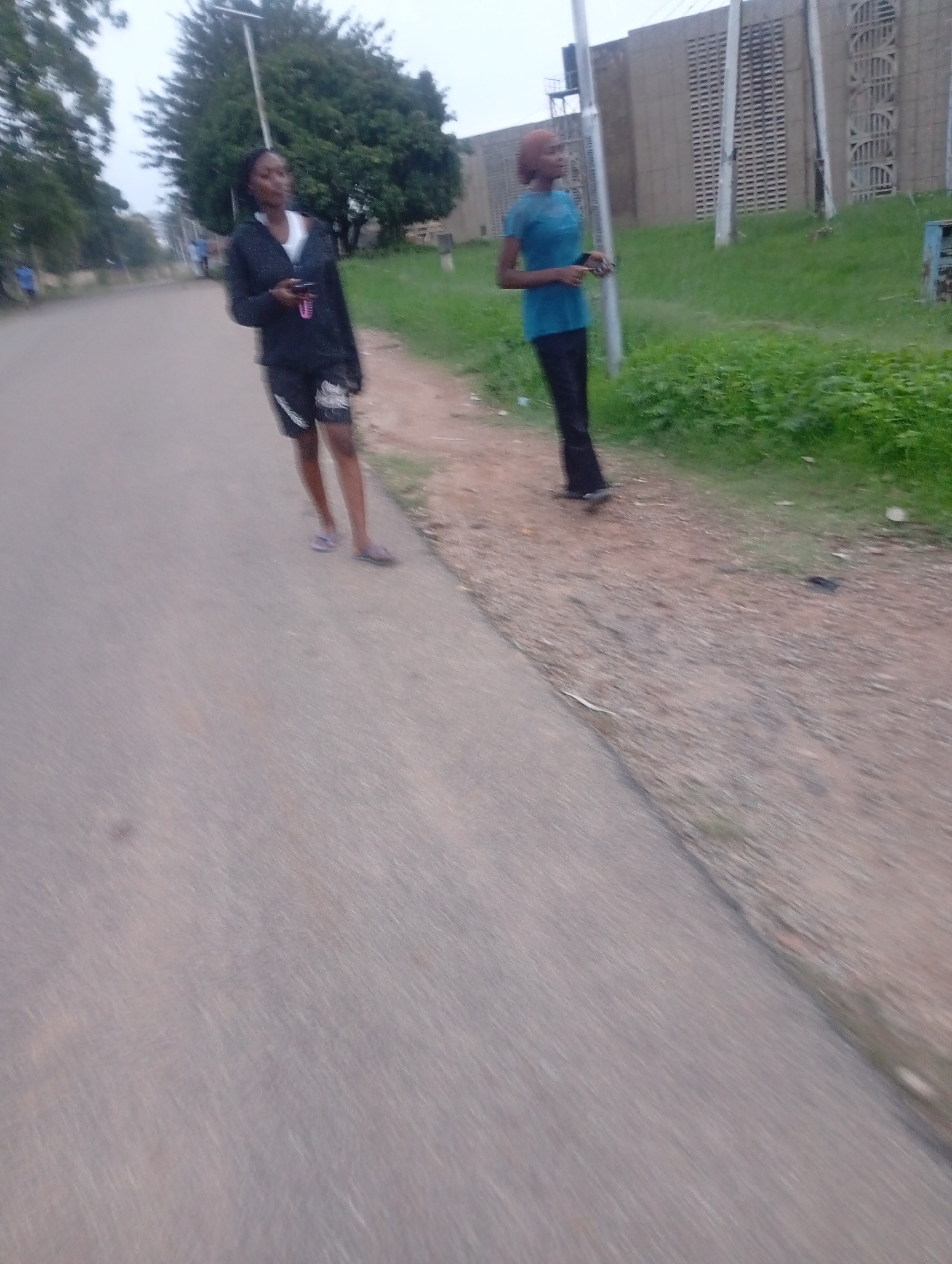 Two young women are standing on a paved road in Jos, Nigeria. The woman on the left is wearing a black jacket and shorts, with white and black athletic shoes. The woman on the right is wearing a blue shirt and black pants. The background shows a grassy embankment with a building composed of concrete blocks and a metal tower. The sky is overcast.