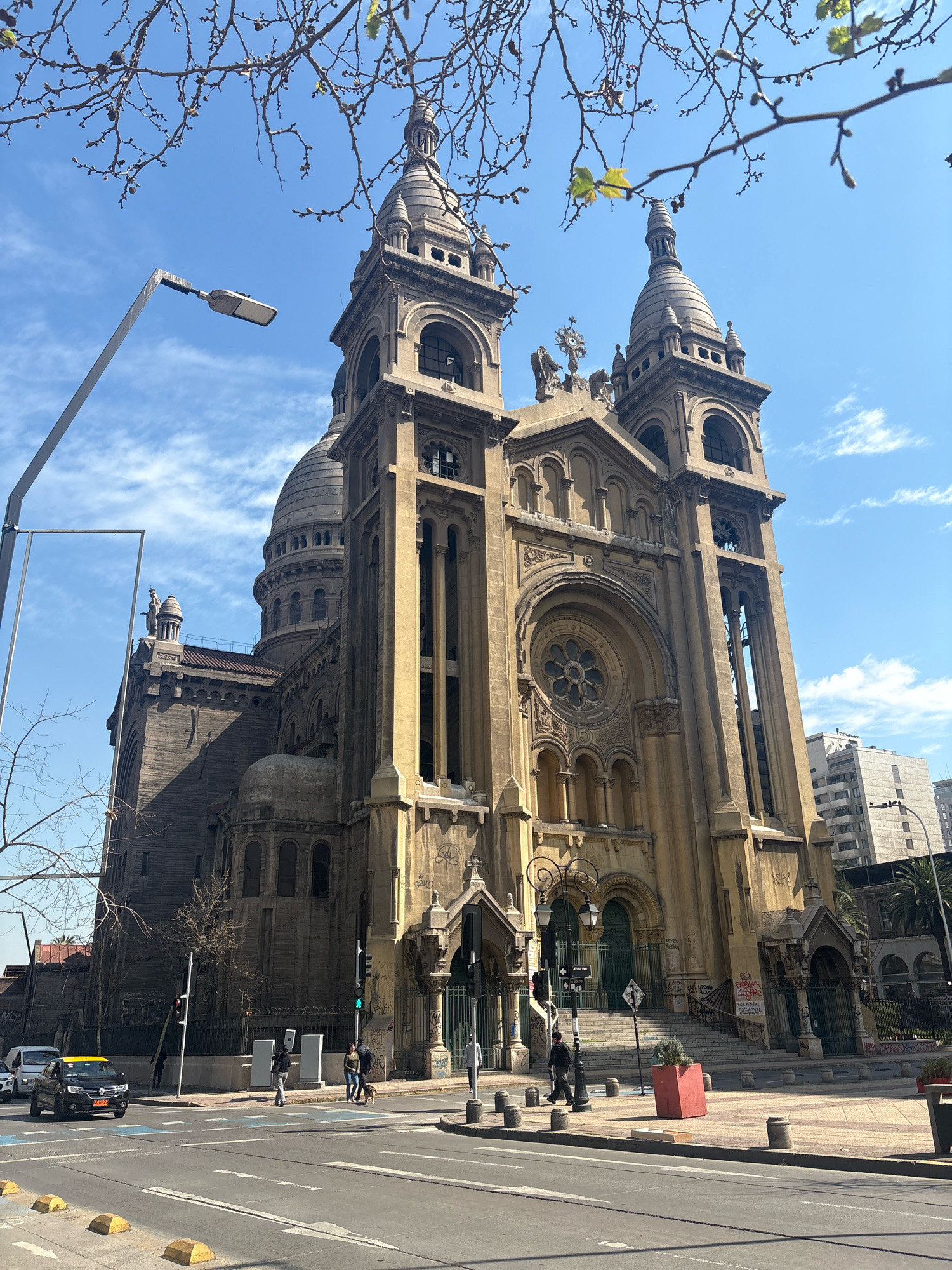 The image shows the Metropolitan Cathedral of Santiago, Chile, on a sunny day with a clear blue sky and scattered white clouds. The cathedral is a large, imposing stone building with multiple domes and towers, characterized by intricate architectural details, arched windows, and ornate carvings. The facade is primarily a light brown or beige color, showing signs of age and weathering.
In the foreground, a street runs in front of the cathedral, with traffic lanes marked by white lines and blue markings. Several vehicles are visible, including a black taxi with a yellow roof, a white van, and other cars. Pedestrians are crossing the street and walking on the sidewalk. A street light on a tall pole stands prominently on the left side of the image. Trees with bare branches and some emerging leaves are also visible in the foreground and partially obscuring the top of the cathedral.
The area surrounding the cathedral appears to be an urban environment, with modern buildings visible in the background to the right of the cathedral. The overall scene depicts a typical urban streetscape in Santiago, with the historic cathedral as the central and dominant structure.