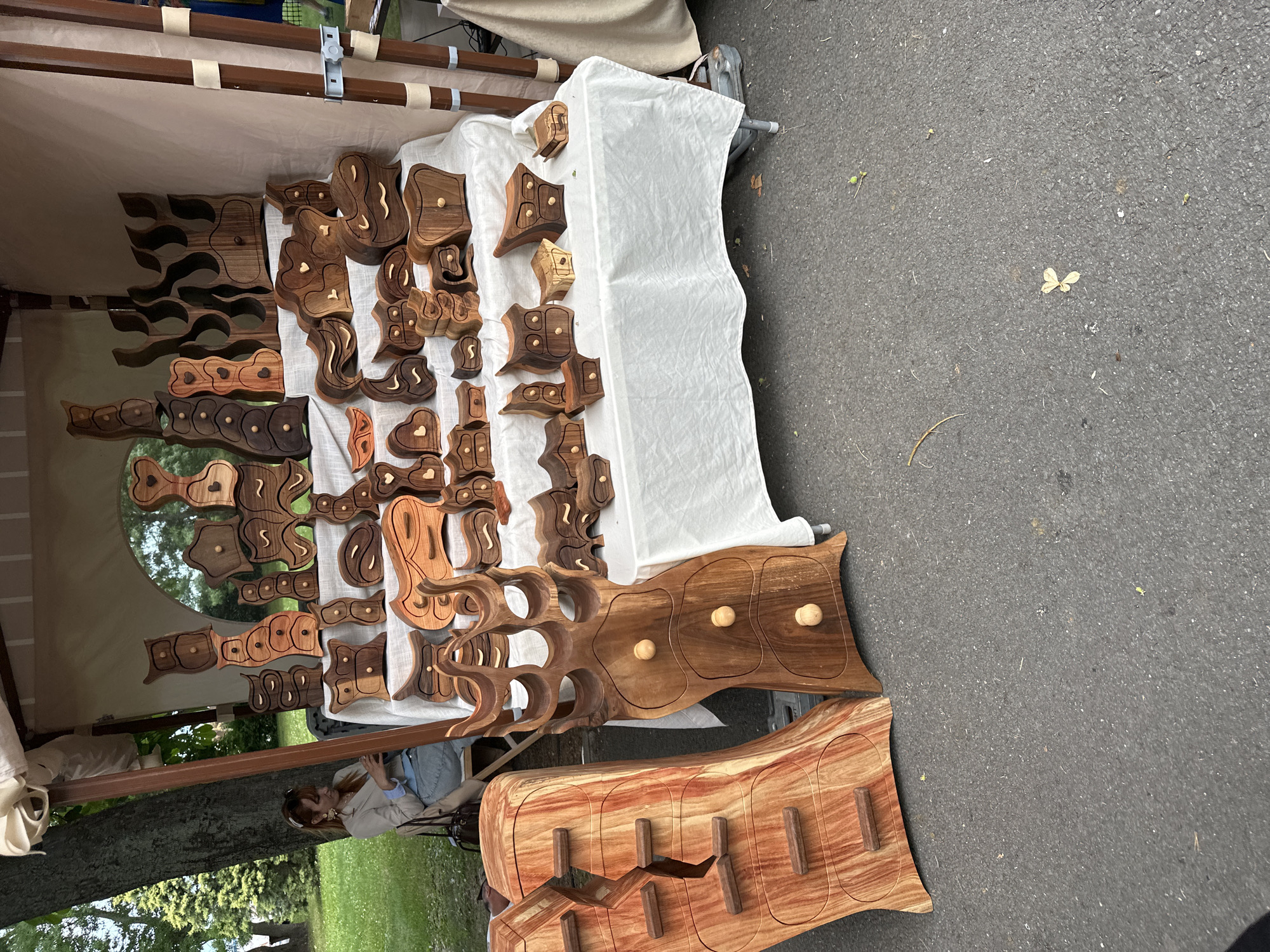 An outdoor market stall, presumably in an unknown city and location, displays numerous handcrafted wooden items. The stall is covered by a beige canvas canopy supported by vertical wooden poles, one of which is visible on the right. A woman with reddish-brown hair, wearing a light-colored jacket over a blue shirt and jeans, sits on a chair in the left background, partially obscured by a tree trunk, looking down at an object in her hands. The background beyond her reveals green grass and more trees.
In the midground, a rectangular table draped with a white tablecloth holds a dense arrangement of unique, organically shaped wooden pieces. These items primarily consist of small to medium-sized drawer units, boxes, and decorative sculptures crafted from various wood types, displaying different natural grain patterns and tones ranging from light tan to dark reddish-brown. Many pieces feature curvilinear contours, abstract forms resembling owls, hearts, or waves, and multiple small drawers with integrated handles or small knobs. Some larger pieces in the back appear to be decorative shelving or wine racks with irregular openings.
In the immediate foreground, resting on a dark gray asphalt surface, are two large, freestanding wooden drawer units. The unit on the left is lighter, reddish-brown, with prominent horizontal grain, designed as two halves with multiple small, rectangular drawers. The unit on the right is darker brown, shaped with an undulating outline, and features three drawers, each with a single spherical light-colored wooden knob. The asphalt ground has scattered small debris and a few light-colored leaves.