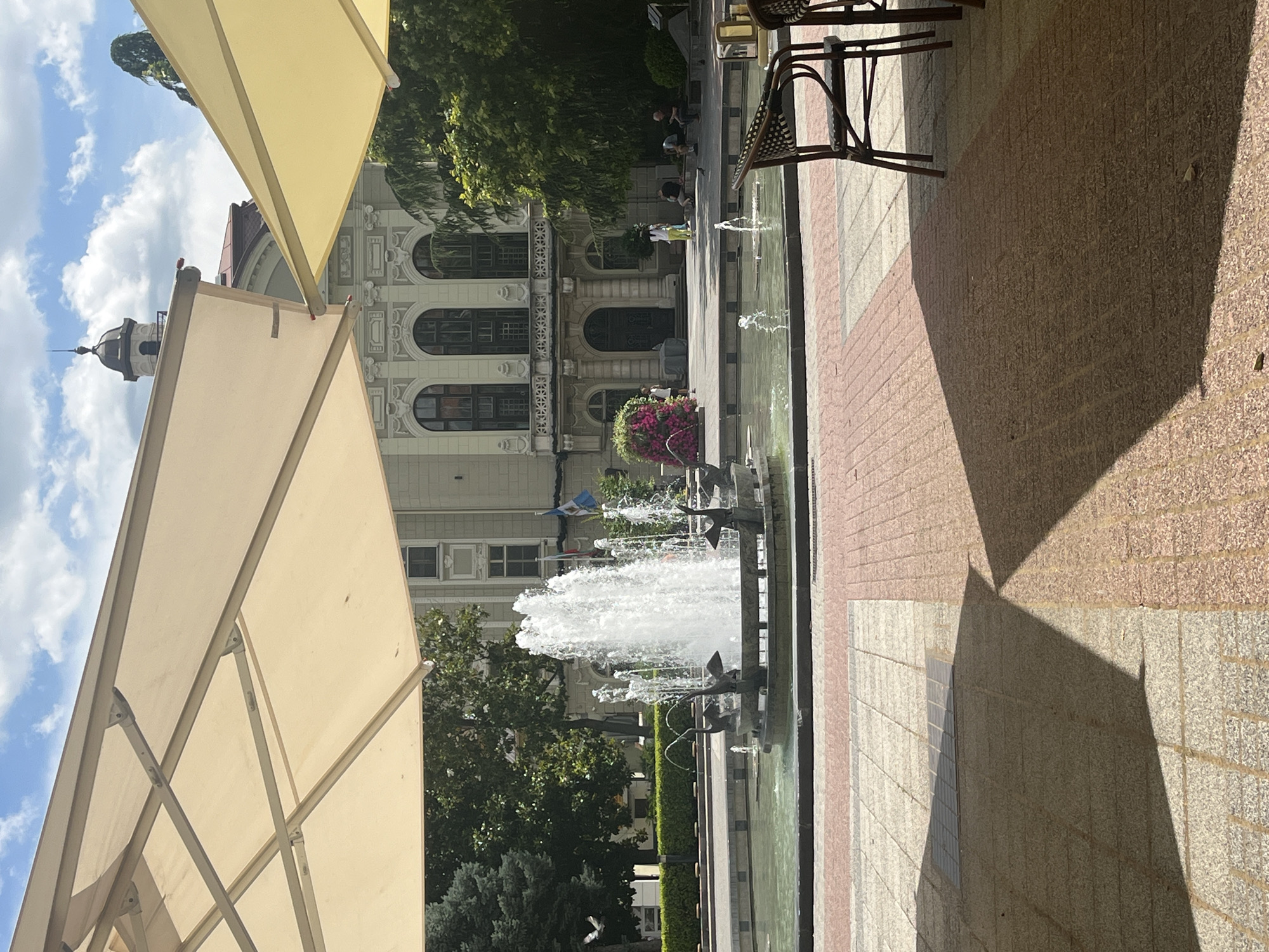 An outdoor scene is captured from the perspective of a cafe, with a large umbrella casting a shadow over the foreground. In the middle ground, a circular fountain with multiple jets of water is visible, adorned with decorative swan sculptures at its base. Beyond the fountain, a paved plaza features several people sitting on benches and a woman standing near a vibrant pink floral arrangement. A grand building with arched windows and ornate architectural details stands in the background, partially obscured by lush green trees. The sky above is a mix of blue with scattered white clouds. This scene is located in the central part of Plovdiv, Bulgaria, likely near the main pedestrian street or a central square.