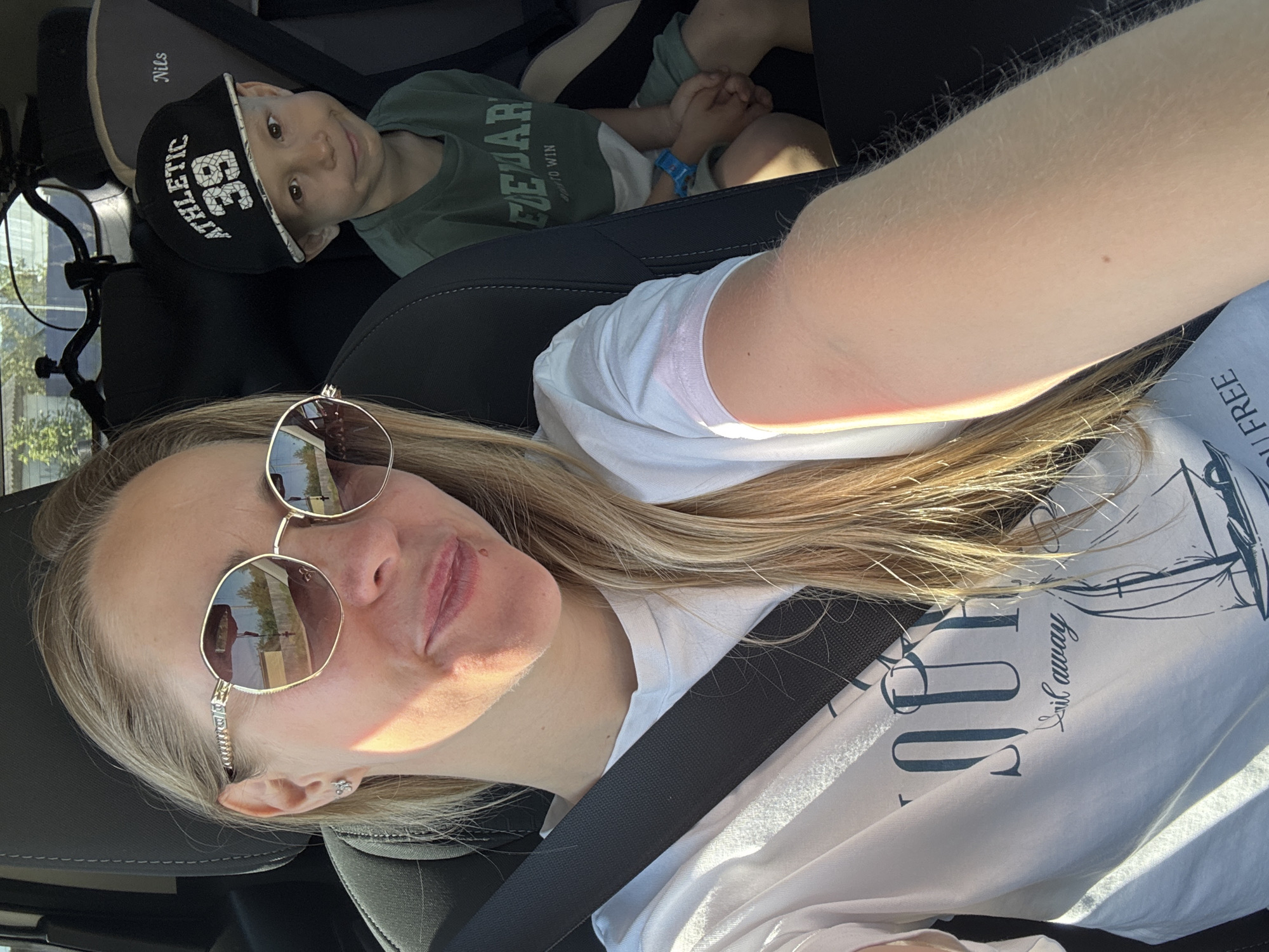 A woman wearing sunglasses and a white t-shirt is in the driver's seat of a car, secured by a seatbelt. Behind her, a young boy in a green shirt and a baseball cap sits in the back seat. The location is likely in Chaikovsky, Russia, based on reflections of buildings in the sunglasses.