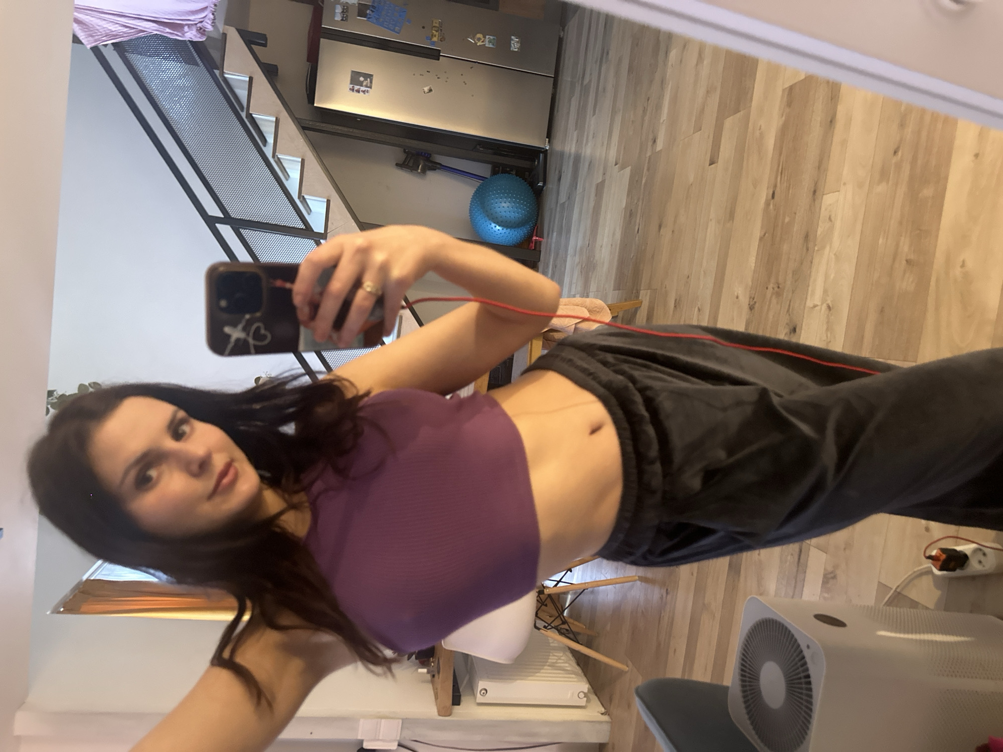 A young woman with long, dark hair and brown eyes is taking a mirror selfie. She is wearing a purple tank top and dark grey sweatpants. Her right hand is holding her phone, and her left arm is bent at the elbow. She is standing in what appears to be a home environment.
Behind her, a staircase with metal railings is visible, leading up to a landing. Further back, there is a stainless steel refrigerator with various items attached to it. A bright blue exercise ball rests on the floor next to the refrigerator. The floor is made of light-colored wood planks. A white air purifier is situated in the foreground, and a power strip with an orange plug is visible on the floor to the right. The overall impression is of a casual, indoor setting, possibly a living area or a multi-purpose room. The lighting suggests it might be daytime.