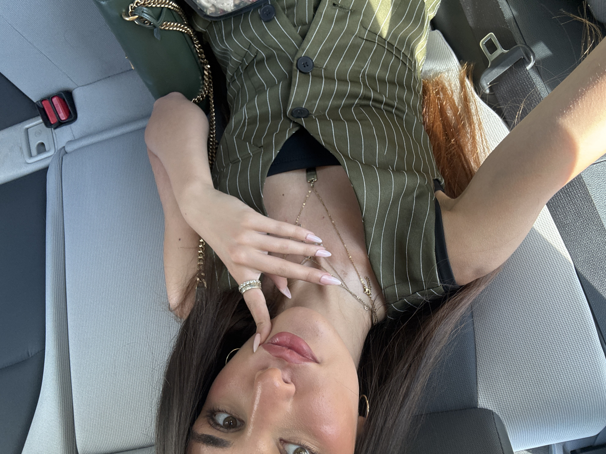 A woman with long, dark brown hair is taking a selfie while seated in the back seat of a car, likely in Aventura, Florida, given the sunlight and typical car interior. She is wearing a green pinstripe vest over a black top and several gold necklaces. Her right hand is raised, touching her chin, and she is wearing rings on her fingers. A green purse with a gold chain strap is visible on her right side. The car seats are gray and black fabric, and a seatbelt is buckled in the background. The lighting suggests it is daytime.