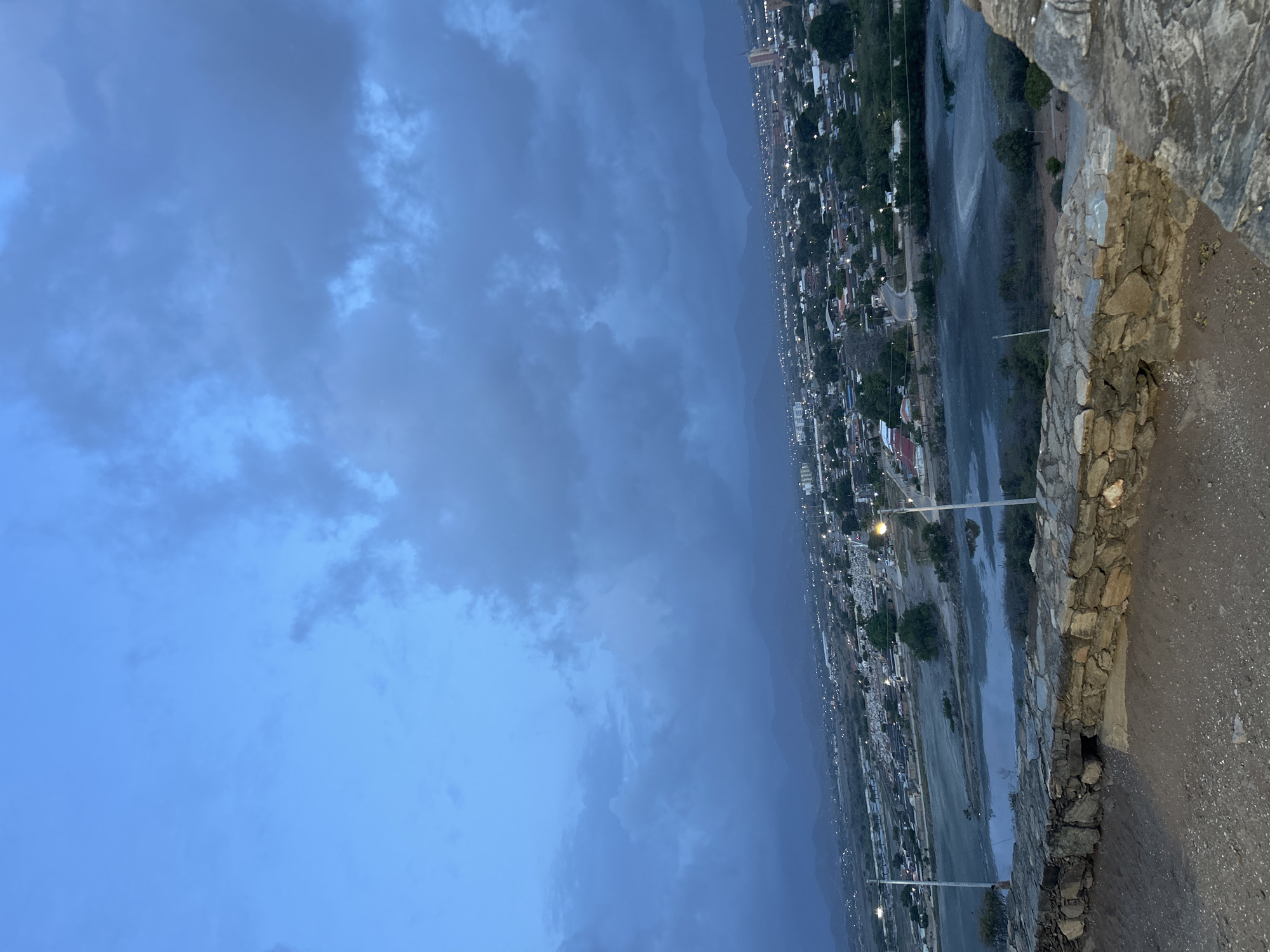 The image captures a panoramic view of the city of Juan Griego, Venezuela, at dusk or early night. The sky is a deep blue transitioning to a darker grey-blue, heavily overcast with dense, dark clouds. A range of mountains forms the distant horizon, silhouetted against the dim light. Below, the urban sprawl of Juan Griego extends, illuminated by scattered streetlights and building lights, appearing as numerous small, warm points of light. In the midground, a large, calm body of water, likely a lagoon or bay, dominates, reflecting the dim sky and appearing murky grey-blue with lighter surface variations. Several roads and additional streetlights are visible within the city grid. The viewpoint is from an elevated position, evidenced by a prominent, rough-hewn stone wall in the immediate foreground, with exposed earth and gravel at its base. Two tall streetlights stand in the midground, illuminating areas near the water. A single, dark human figure is faintly visible standing near the water's edge on the left side of the frame, partially obscured by a streetlight pole. No discernible actions or interactions are taking place.