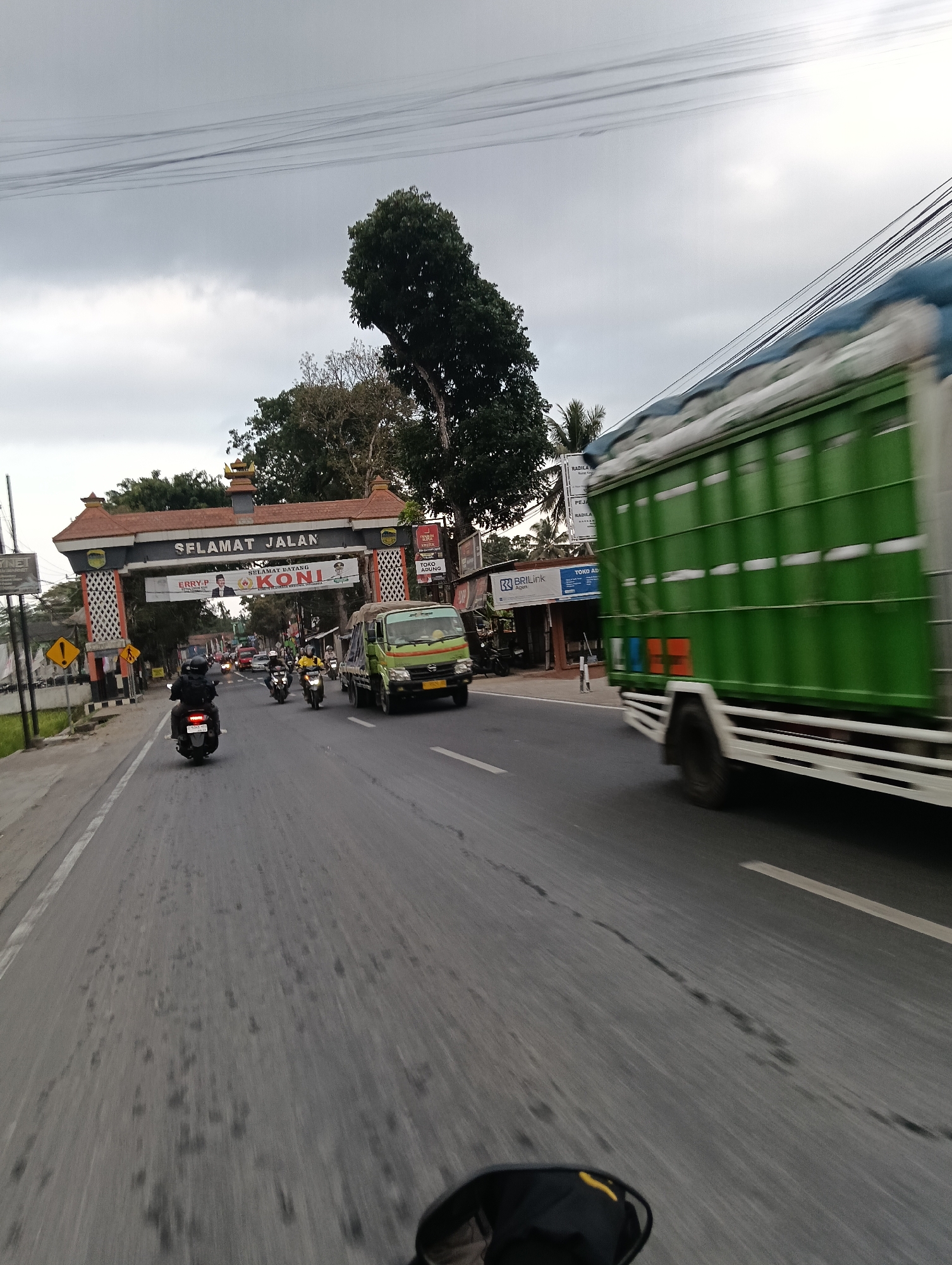 The image depicts a street scene in Cintaraja, Indonesia. A banner over an archway reads "SELAMAT JALAN," and another banner below says "SELAMAT DATANG." Several motorcycles are visible on the road, with one in the foreground and others further ahead. A green truck is in motion on the right side of the frame, heavily blurred due to the speed. The sky is overcast. Buildings line the right side of the street, including what appears to be a "BRIlink" branch. The road surface shows signs of wear and tear, with dark patches and some cracks. The perspective suggests the photo was taken from a moving vehicle.