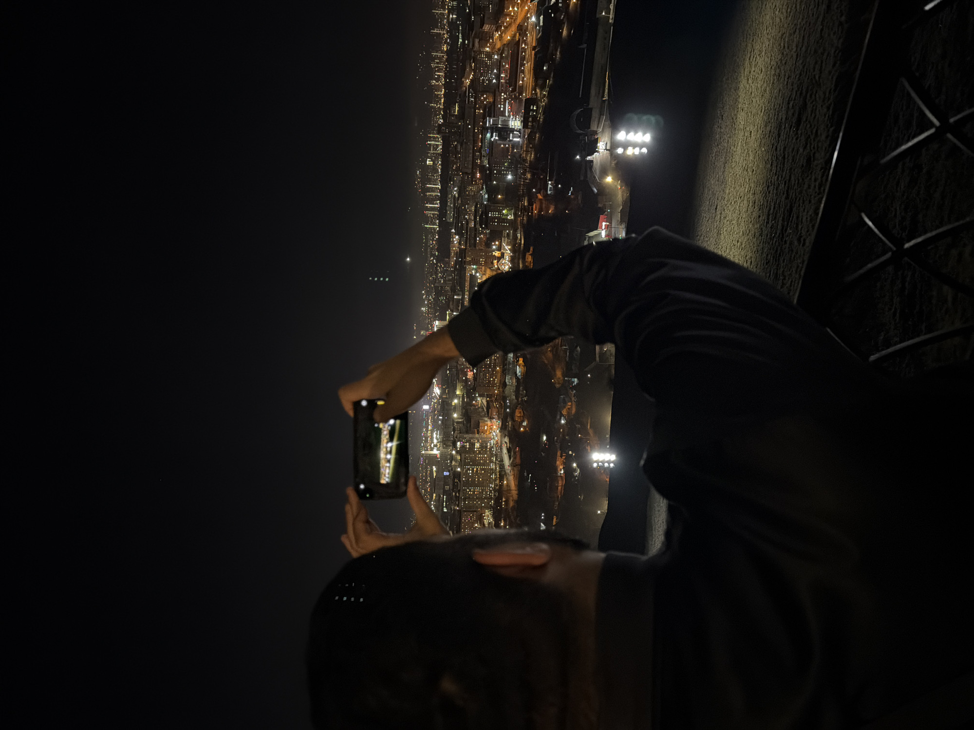 At night, an individual photographs the city of Krasnoyarsk, Russia, from a high vantage point. The person, facing away from the camera, is holding a smartphone up to capture the city skyline. The city, illuminated with numerous lights, stretches out into the distance. To the right of the photographer are bright floodlights and the edge of what appears to be a barrier or fence. The sky above is dark.
