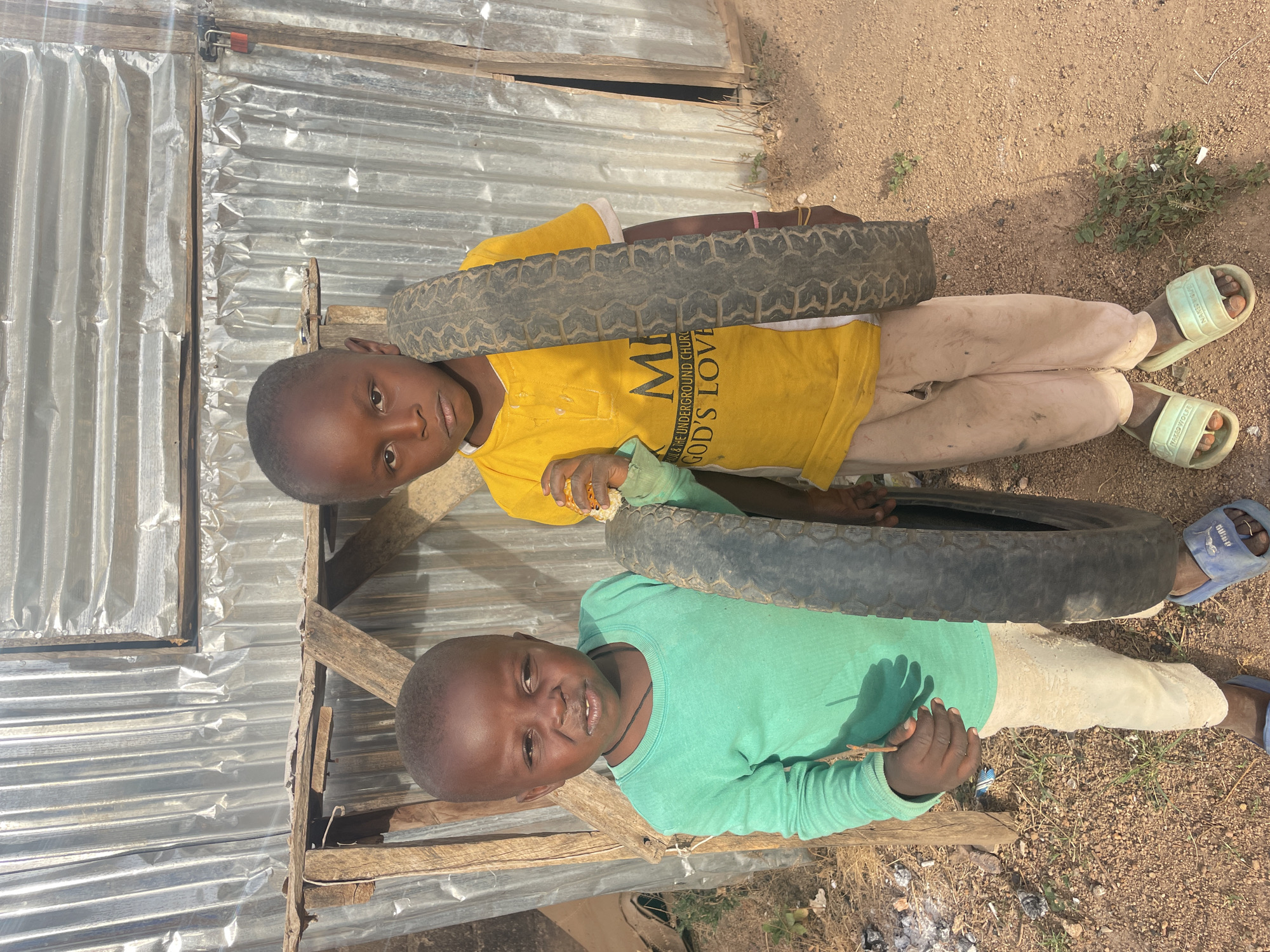 Two young boys are captured in a bright, sunlit outdoor setting in Barakin Lerawi, Nigeria. The image is oriented vertically, showing the boys looking up towards the camera.
The boy on the left is wearing a yellow short-sleeved t-shirt, light-colored long pants, and light green slides. On his t-shirt, partially visible text reads "M... THE UNDERGROUND CHURC... GOD'S LOVE." He holds a small, black, worn tire in front of him. His expression is neutral as he looks directly at the camera.
To his right, the second boy is dressed in a light green long-sleeved shirt, light-colored long pants, and blue slides. He also holds a similar small, black tire. He has a slight smile and looks towards the camera. Both boys have short, dark hair.
The setting is characterized by dry, sandy or dirt ground with sparse vegetation. In the background, there is a structure made of corrugated metal sheets and wooden supports, suggesting a simple building or fence. The bright sunlight indicates it is likely midday or early afternoon, casting clear shadows. The boys appear to be posing, possibly in preparation for or during play with the tires.