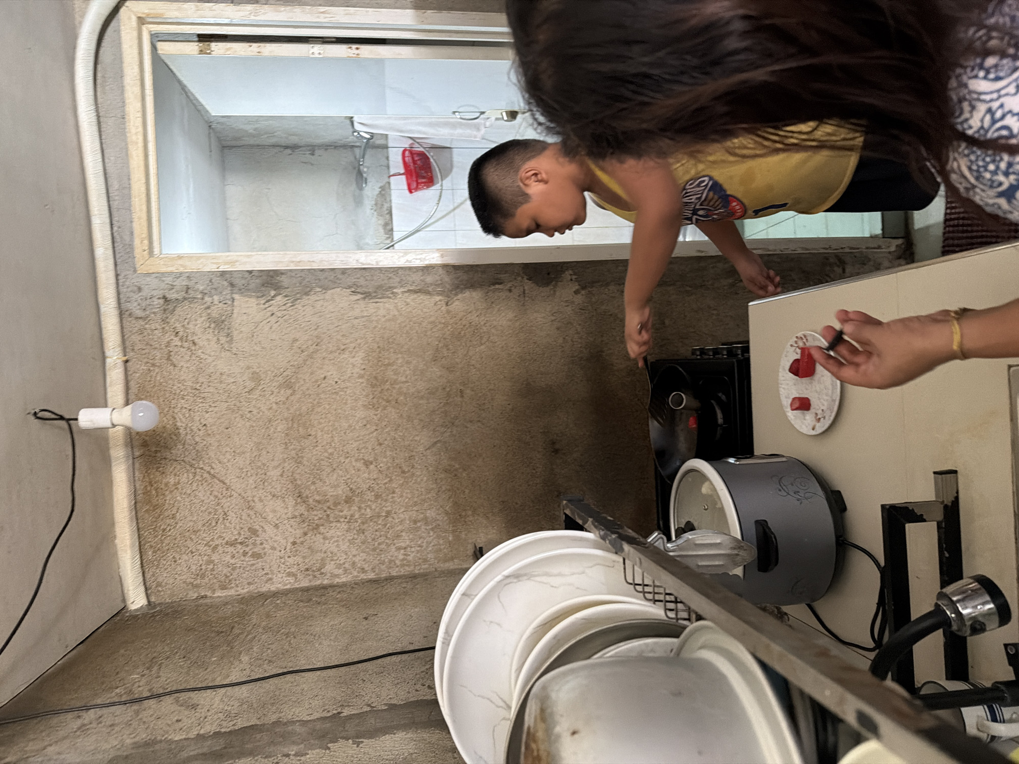 The image depicts a dimly lit kitchen in Davao City, Philippines. A young boy, wearing a yellow tank top with a graphic print, stands near a stove, appearing to be cooking or preparing food. He is looking towards the stove with his right hand extended as if tending to something cooking in a pan. In the foreground, the back of a woman's head and shoulders are visible; she is wearing a patterned top and has a gold bracelet on her left wrist. She is also looking towards the area where the boy is cooking. A plate with pieces of what appear to be fruit or vegetables is placed on the counter.
The kitchen environment is basic and functional. A dish rack filled with white plates is positioned on the left side of the frame. A gray rice cooker sits on the counter. The walls have a rough, unplastered texture, and a bare light bulb hangs from the ceiling, providing the primary illumination. A doorway in the background leads to what seems to be a bathroom, with a shower head and a red plastic basket visible. Electrical wires are strung along the walls and ceiling. The overall impression is of a lived-in, utilitarian space.