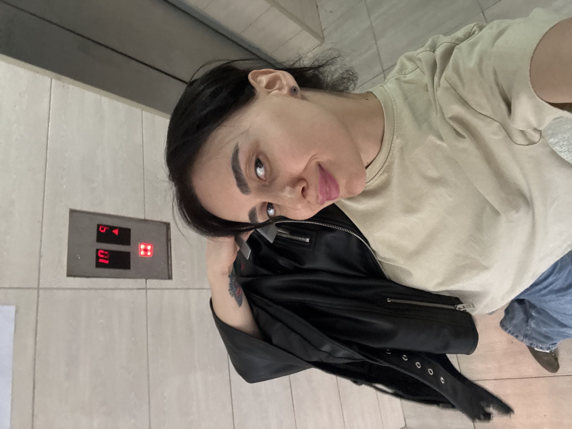 A selfie taken indoors shows a fair-skinned individual with dark hair and arched eyebrows. The person is wearing a light beige t-shirt, blue jeans, and a black leather jacket is draped over their arm. The background suggests an elevator, with a control panel displaying the number "10" and an upwards arrow illuminated in red. The surrounding walls appear to be tiled. The location cannot be confirmed as Tbilisi, Georgia, without additional information.