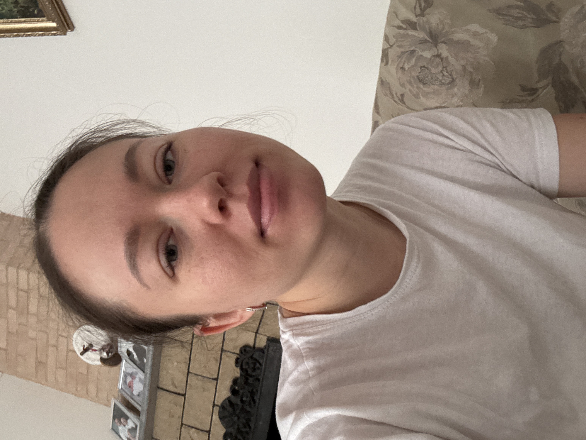 A young woman with brown hair pulled back is wearing a white t-shirt. She is indoors, with a floral pattern pillow behind her to the right, and a brick wall with a fireplace and framed photos to the left. The location in the city of Ferma, Russia is unknown.