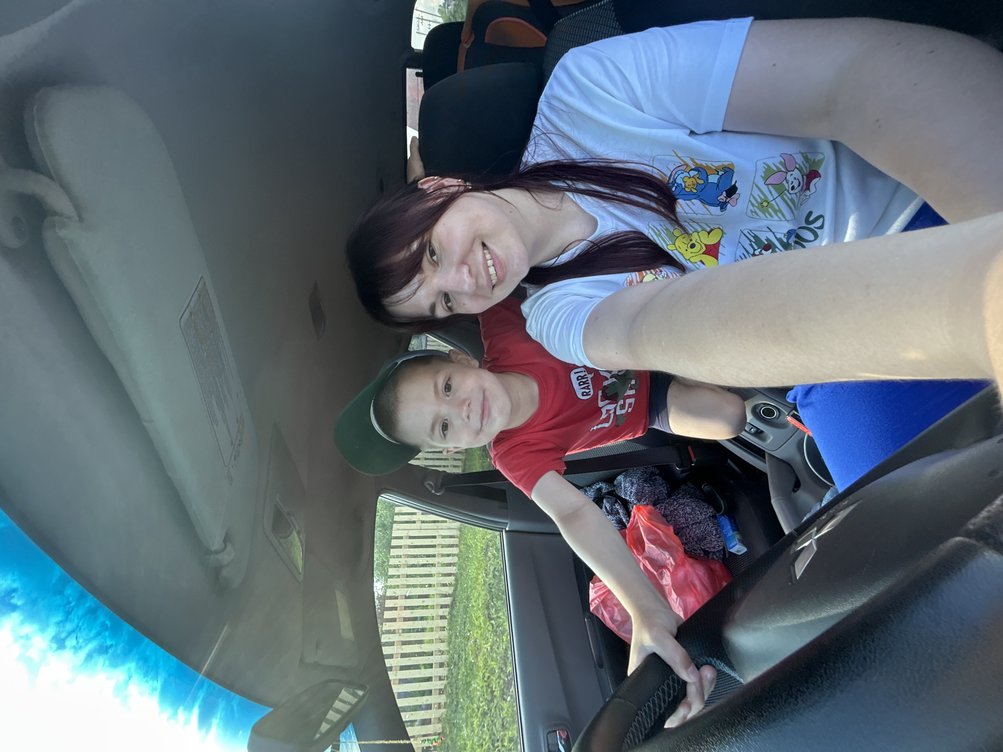 A mother and child are in the front seats of a car. The mother is on the right side of the frame, smiling at the camera, wearing a white t-shirt with Winnie the Pooh characters. The child, on the left, is wearing a red t-shirt and a green baseball cap backwards. He is holding the steering wheel and looking at the camera. The car interior is visible, including the dashboard, steering wheel, and seats. Outside the car window, a wooden fence and green grass are visible. The scene is illuminated by natural sunlight coming through the windshield and sky. The context of the location in the city of Фоки, Russia, is not discernible from the visual information in the image.