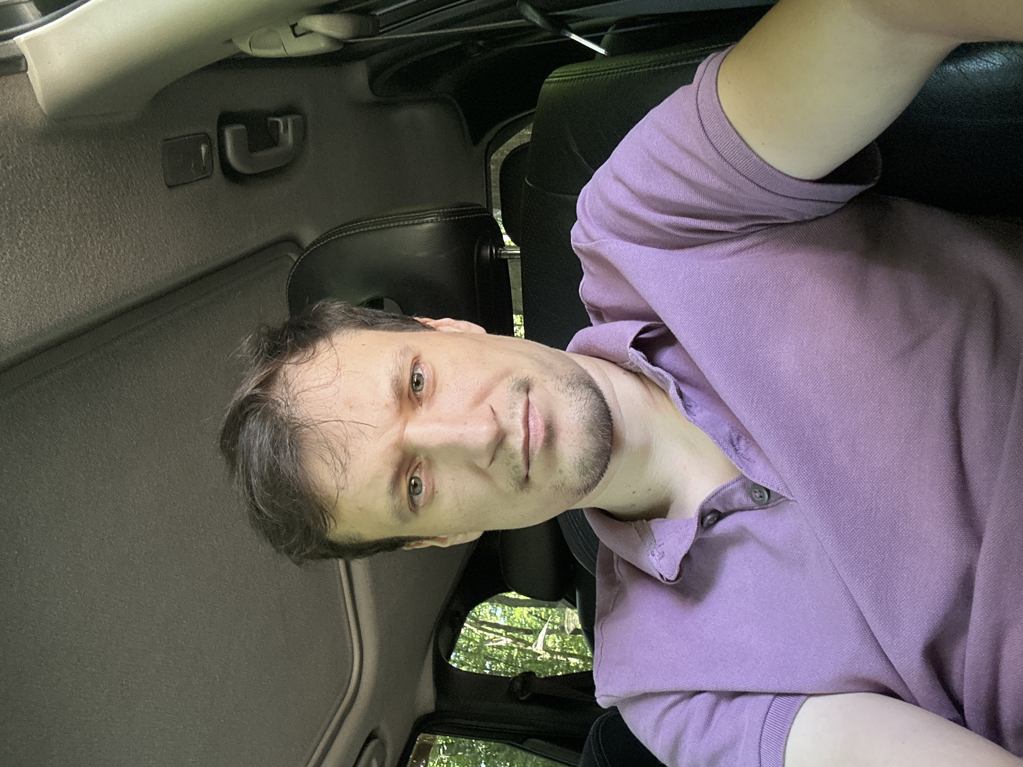 A medium shot shows a man from the chest up, sitting in the driver's seat of a car, likely in Shatava, Ukraine. He has light skin, short brown hair, a light beard, and green eyes. He is wearing a purple polo shirt. The car's interior is visible, including the black leather seats, the roof lining with a sunroof, and a portion of the dashboard. Through the windows, trees and greenery are visible, suggesting an outdoor, possibly rural, setting. The man is looking directly at the camera, his expression neutral. The lighting is natural, coming from the front and sides.