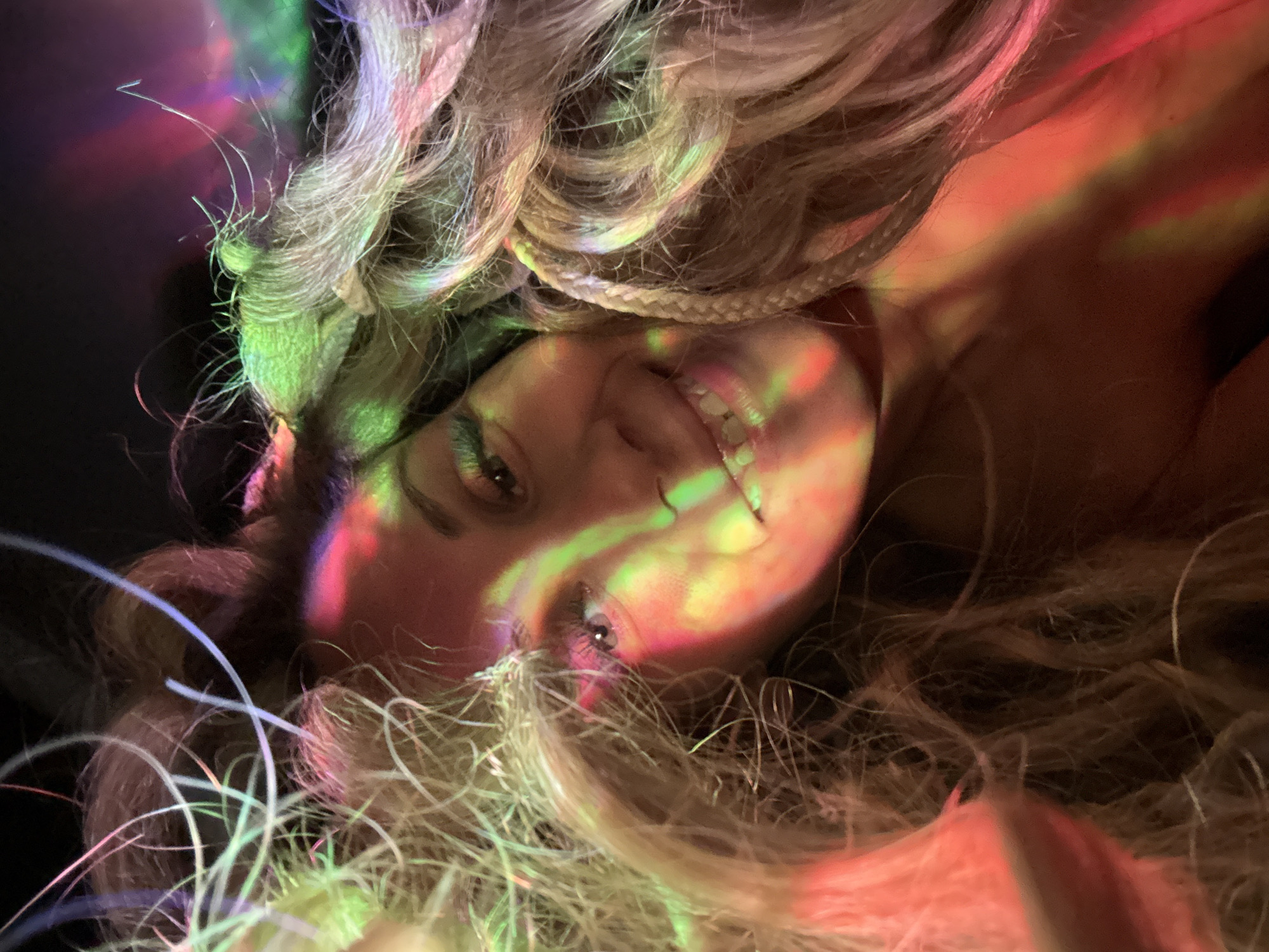 A close-up, high-angle portrait of a woman with long, wavy, blonde hair illuminated by multicolored lights (green, red, and pink) casting shadows on her face. She is smiling, revealing her teeth, and one braid is visible amongst her hair. Her eyes are open and visible, partially obscured by strands of hair. A dark background surrounds the subject and portions of her hair. There is no evidence indicating the location of Rusanovo, Russia, or any other specific place.