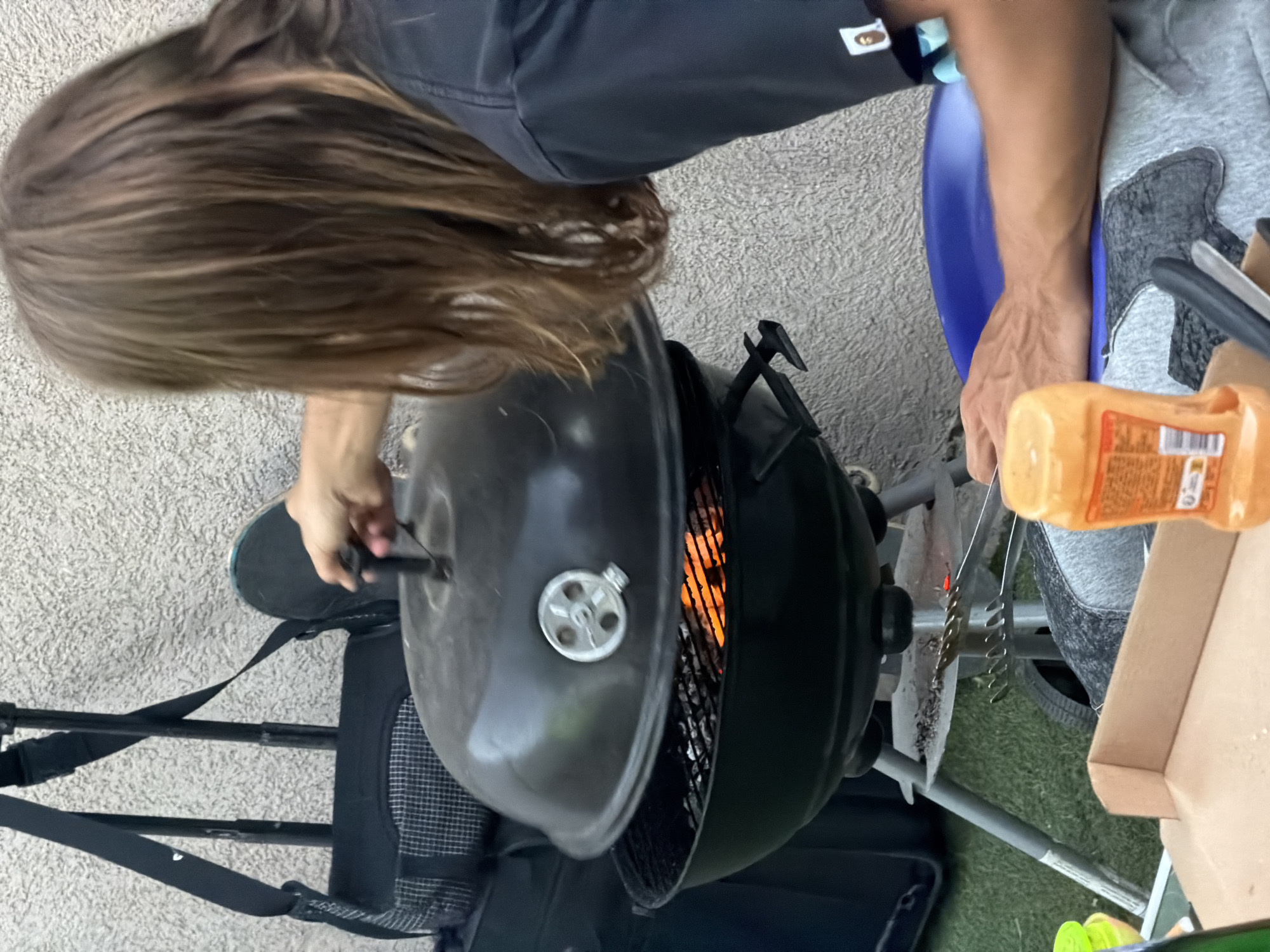In Villeurbanne, France, a person with long, light brown hair is attending to a charcoal grill, partially covering it with a black lid. The grill contains glowing embers, indicating it is hot. The person is wearing a black t-shirt with a small graphic on the sleeve. In the foreground, a bottle of orange sauce is visible, along with a pizza box. Luggage and a backpack are present in the background. The ground appears to be a mix of grass and pavement.