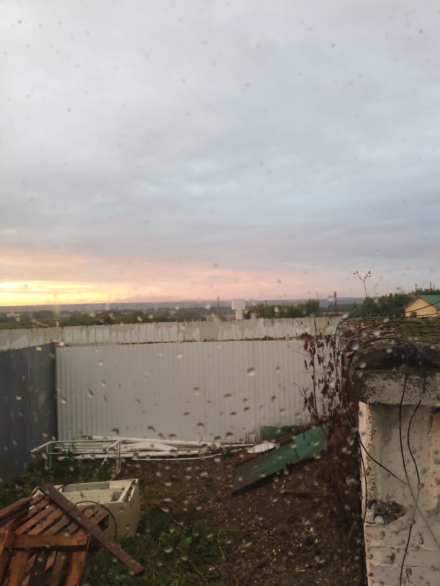 The image depicts a view through a window with raindrops on the glass, showing an outdoor scene in Platoshino, Russia. The time of day appears to be either sunrise or sunset, with a hazy sky displaying muted orange and pink hues near the horizon, transitioning to a grey overcast sky above.
In the foreground, the view is partially obscured by what appears to be a section of a damaged building or structure on the right, made of concrete blocks and painted white, with dark wires or vines attached. Some debris, including what looks like a broken wooden pallet and a discarded white appliance, is visible on the ground in the lower left quadrant.
Beyond this, a corrugated metal fence stretches across the mid-ground, separating the immediate area from a landscape beyond. The landscape features a line of green trees, and further in the distance, what seem to be scattered buildings, including a small yellow building with a green roof on the right side. The ground outside the fence is covered in dirt and some sparse vegetation. The overall impression is one of a somewhat unkempt or rural outskirts area.