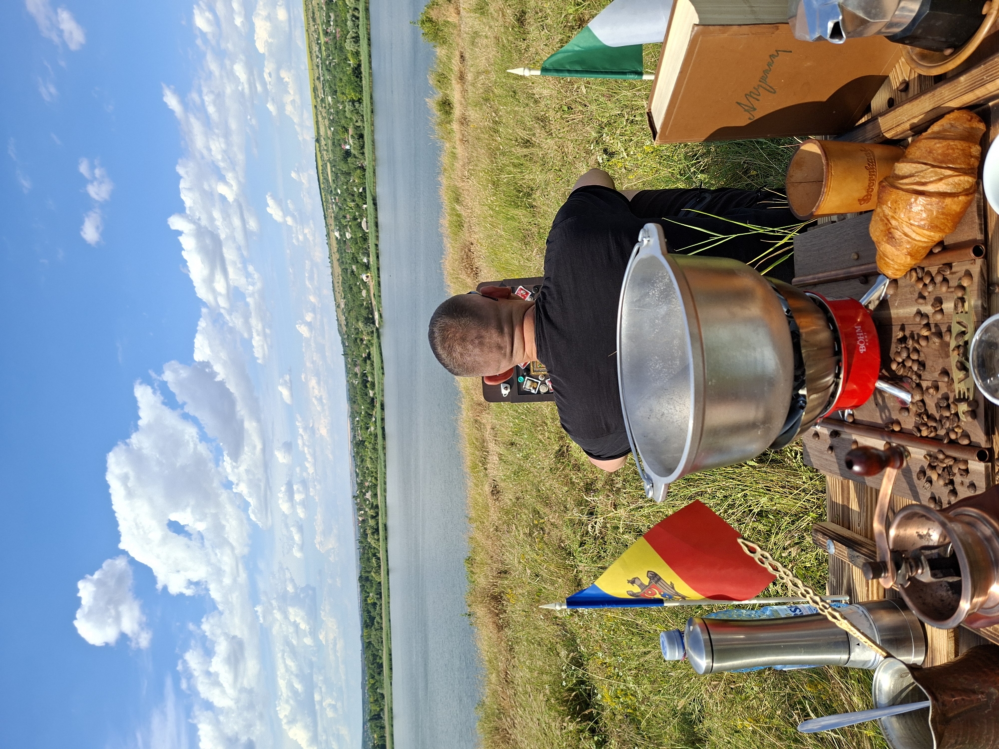 The image depicts a rear view of a man seated outdoors, facing a body of water. The man is wearing a black t-shirt. In the foreground, a wooden table is laden with various items, including a red camping stove with a pot on top, a manual coffee grinder, coffee beans, a croissant, a book, a Moka pot, a stainless steel water bottle, and two small flags – one of Moldova and one of Italy. The environment is grassy and rural, with a large body of water stretching into the distance. In the background, rolling hills covered with trees and scattered houses suggest a rural or semi-rural setting. The sky is blue with scattered white clouds. Based on the presence of the Moldovan flag, the scene is likely located in Oxentea, Moldova.