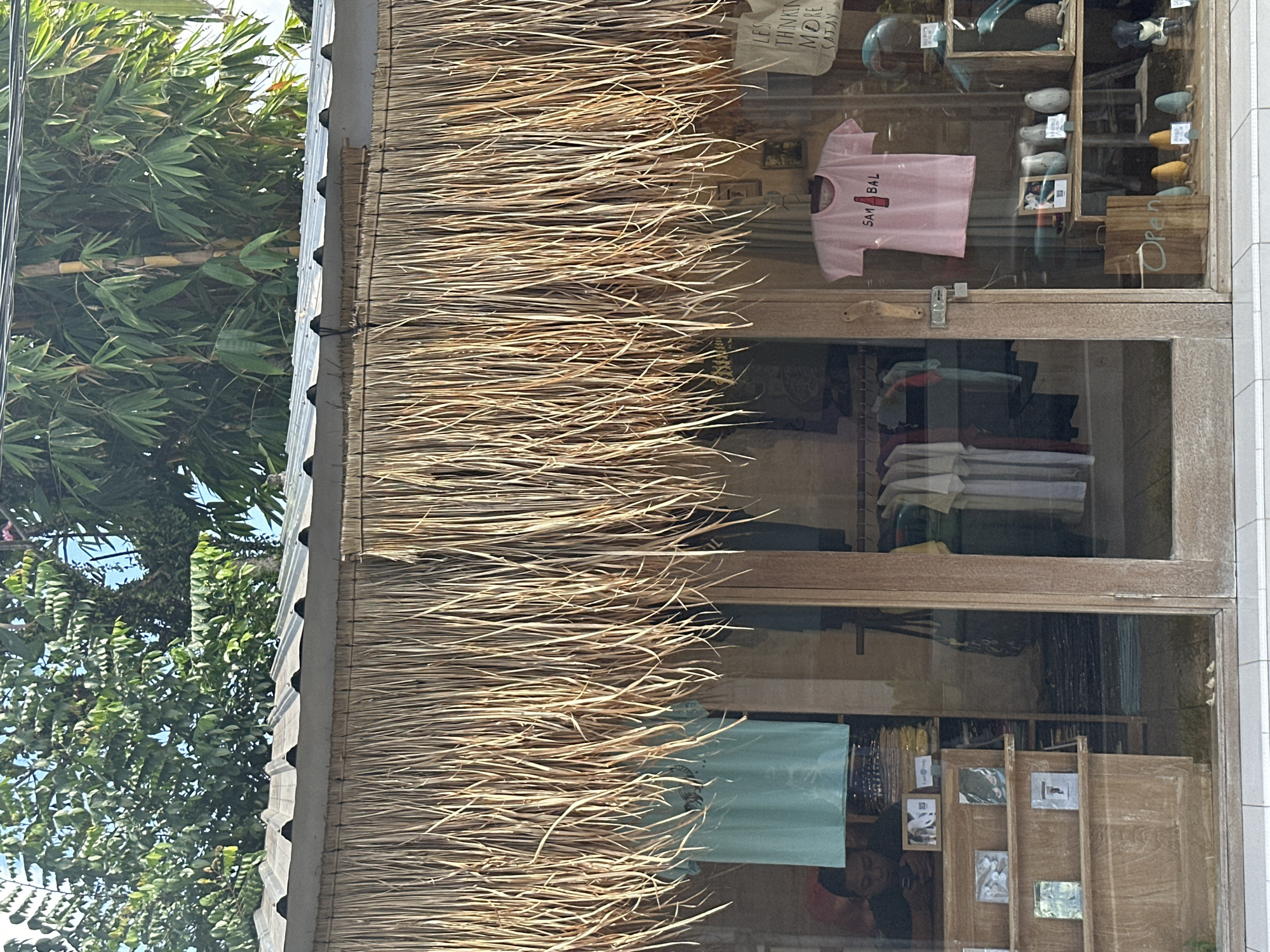 A retail storefront with a thatched roof overhang in Ubud, Indonesia, displays merchandise through its glass facade. A person is visible in the left section of the store, looking at a mobile phone. The store's interior features wooden shelves stocked with various items, including what appear to be books or art prints, as well as clothing racks with t-shirts and dresses. A pink t-shirt with the text "SAM BAL" and an illustration of a bottle is prominently displayed on the right side of the storefront. Various decorative items, including small figurines and what seem to be small ceramic objects, are also arranged on shelves. A wooden sign that reads "Open" is placed on the floor in front of the entrance. The background shows lush green trees and bamboo stalks.