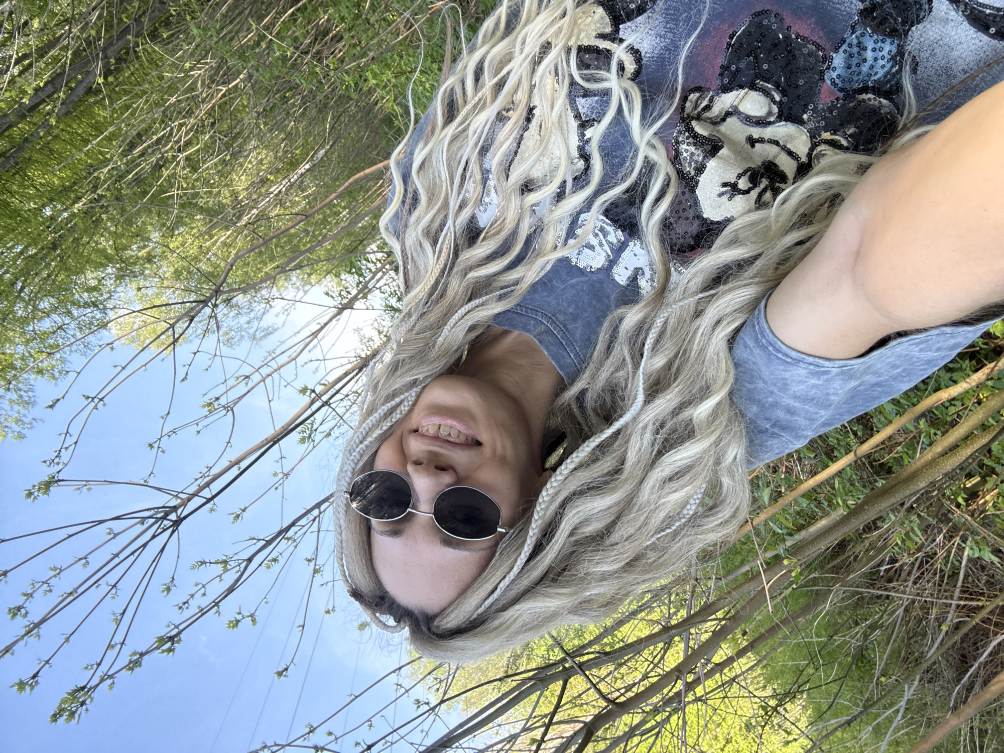 A young woman poses for a selfie outdoors. She has long, blonde hair with small braids interspersed. She is wearing round, dark sunglasses and a grey t-shirt with a dark floral or sequined design. The background shows trees and branches with emerging green leaves against a blue sky, suggesting it is spring. The location is likely a park or wooded area. The photograph's orientation is rotated. The subject and surroundings are consistent with a natural environment that might be found in a place like Rusanovo, Russia.
