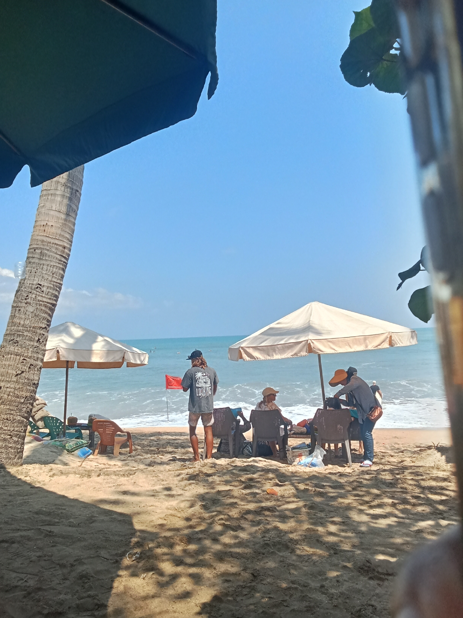 A serene beach scene unfolds in Kuta, Indonesia, under a clear blue sky. A tall palm tree dominates the left side of the frame, its rough trunk casting a shadow on the sandy shore. In the middle ground, two beach umbrellas provide shade, one a pale beige, the other a darker shade of green. Beneath the beige umbrella, a group of people are gathered around a table. One person, wearing a straw hat and a light-colored shirt, sits in a chair. Two other individuals, one with a dark baseball cap and the other with a dark hat, are standing and appear to be interacting with the person seated. A red flag stands planted in the sand near the water's edge, signaling the presence of waves. To the right, the ocean stretches to the horizon, its gentle waves lapping the shore. The sand is a light beige, with some areas appearing darker due to shadows from nearby foliage, which is partially visible in the foreground and right edge of the image, suggesting a natural canopy overhead. The overall atmosphere is one of relaxation and leisure, typical of a sunny day at the beach.