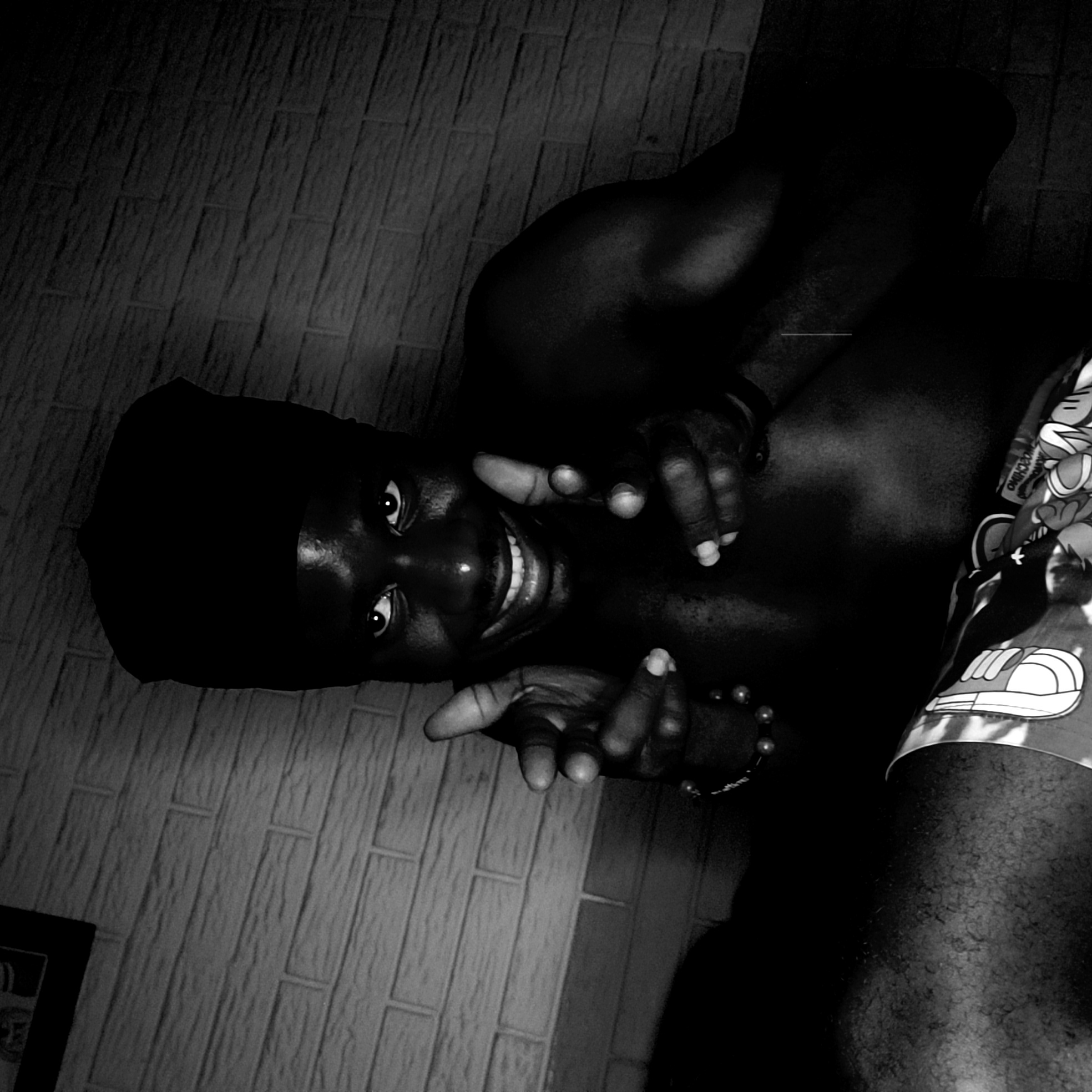 A high-contrast black and white photograph captures a person with dark skin looking directly at the camera with a wide, open-mouthed smile, showing teeth, and expressive, wide eyes. The individual, identified as male, is positioned at an angle and wears a dark head covering and patterned shorts featuring cartoon-style graphics, including a sneaker and what appears to be mirrored text that could be part of "MOSCHINO."
Both of the subject's hands are raised and extended towards the viewer, with fingers slightly curled in an inviting or playful gesture. A beaded bracelet is visible on his left wrist. The lighting is dramatic, creating deep shadows that emphasize his facial features and the muscular contours of his shoulders and arms, while large parts of the image remain dark. The background consists of a dark, textured tiled surface with a rectangular pattern, providing a stark visual contrast. The overall impression is engaging and friendly, set indoors in Narayi, Nigeria.