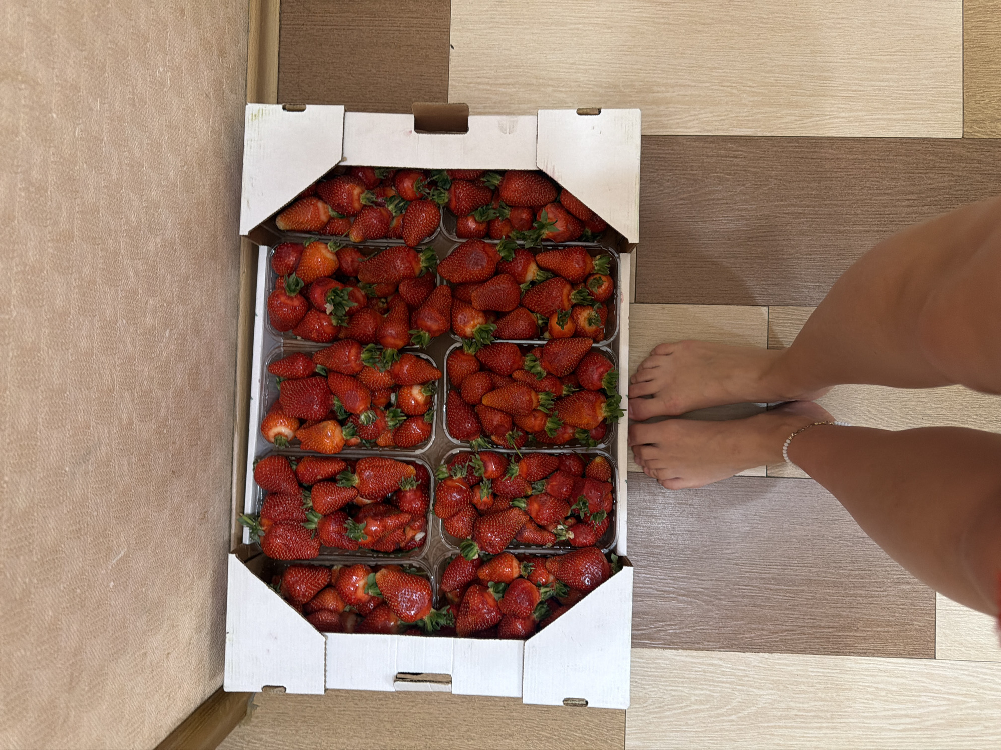 A top-down shot shows a rectangular white cardboard crate filled with fresh strawberries, likely destined for consumption or sale. The crate is divided into compartments, with each compartment holding a clear plastic clamshell container filled with the ripe red fruit. The strawberries fill the container so the container cannot be seen clearly.
The crate rests on a floor composed of light and dark brown planks arranged horizontally and running the length of the floor. To the right of the crate, a pair of bare feet is visible, with a glimpse of a leg extending upwards, including an ankle bracelet. The setting appears to be indoors. The photograph was taken in Moscow, Russia.